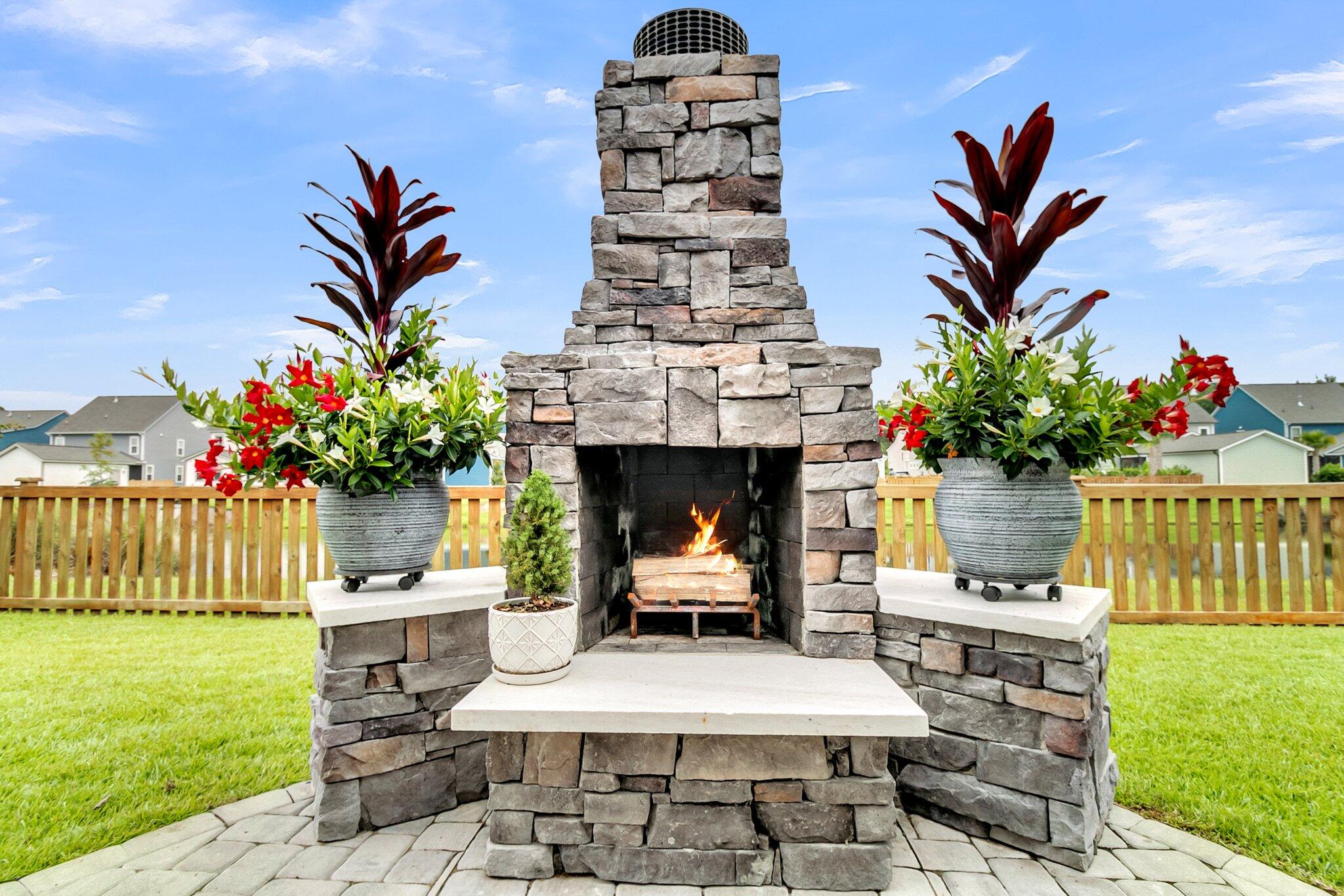 Stone outdoor fireplace burning on a paved patio, flanked by flower-filled planters.
