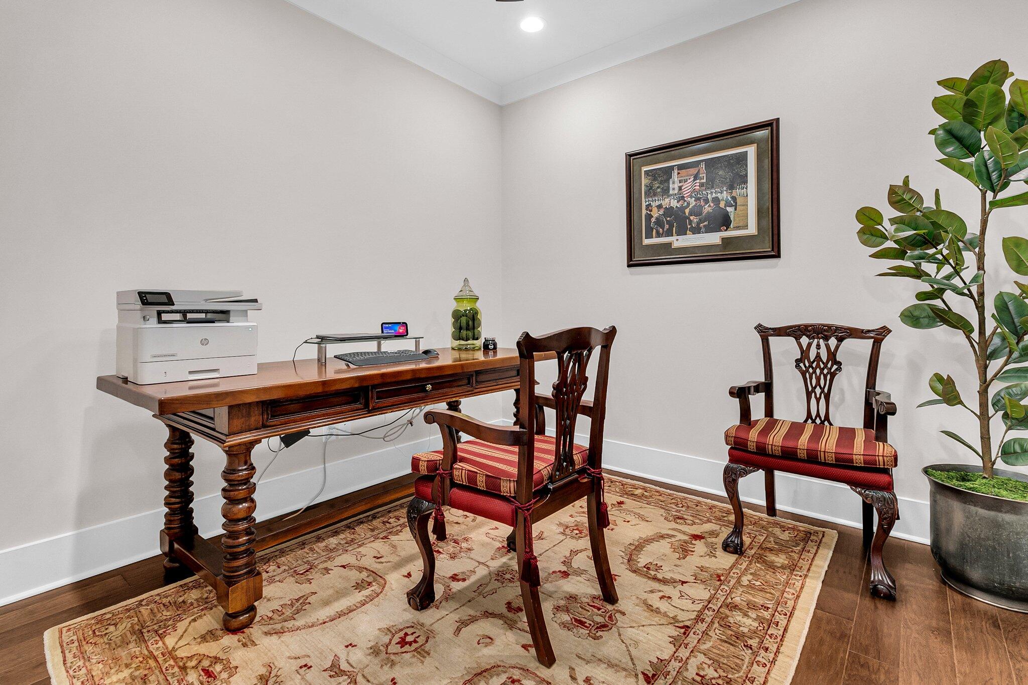 Home office with a carved wood desk, red-cushioned chairs, patterned rug, and tall potted plant.