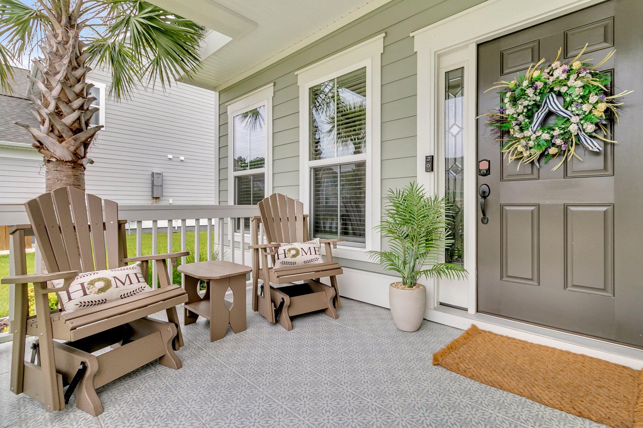 Covered front porch with a wreath-decorated door, patterned tile, potted palms, and two wooden glider benches.