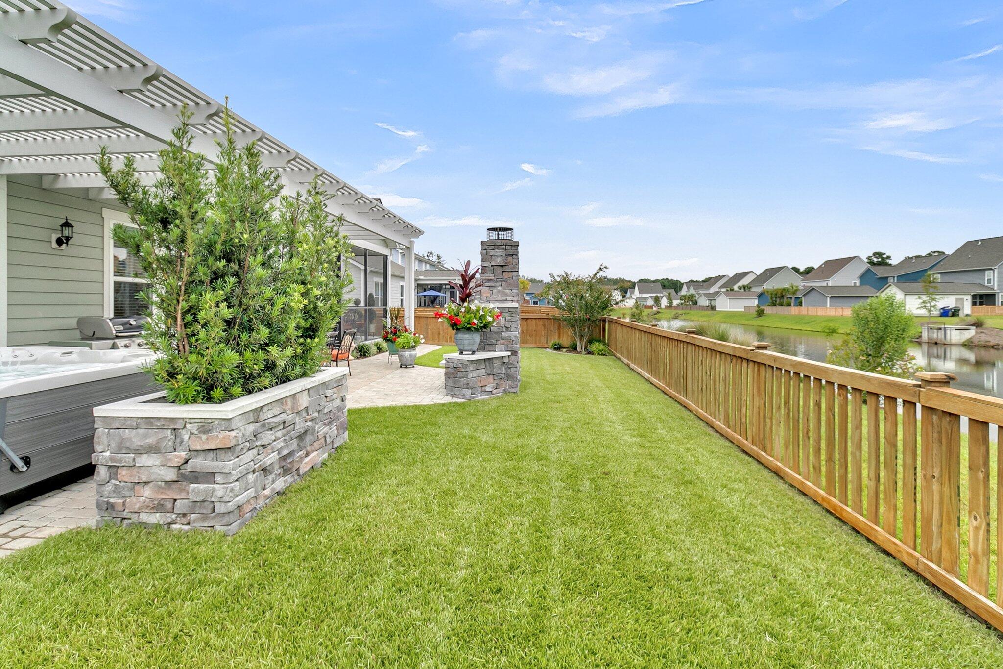 Backyard with a hot tub, stone patio fireplace, fenced lawn, and pond view.