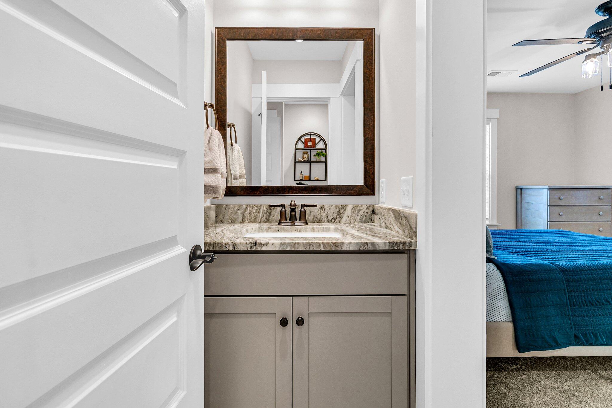 Bathroom vanity with a veined stone counter and framed mirror beside a bedroom with a blue bedspread.
