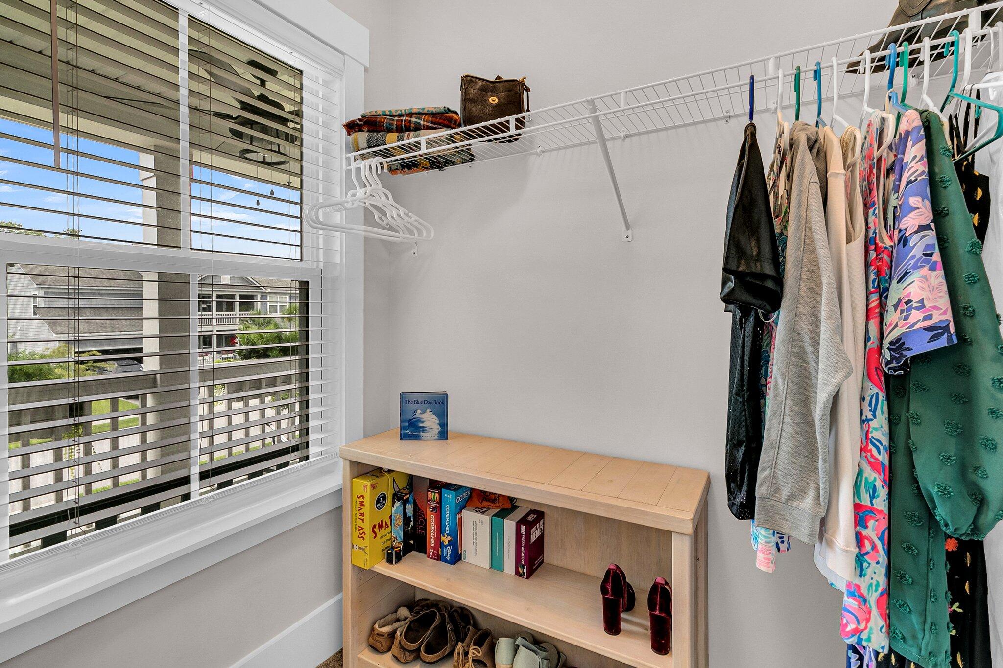 Walk-in closet with hanging clothes, a window with blinds, and a bookcase for shoes and books.