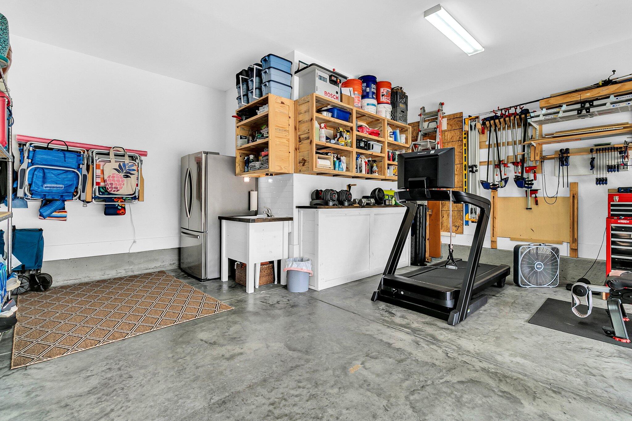 Organized garage with wooden storage shelves, a treadmill, refrigerator, and wall-mounted tools.