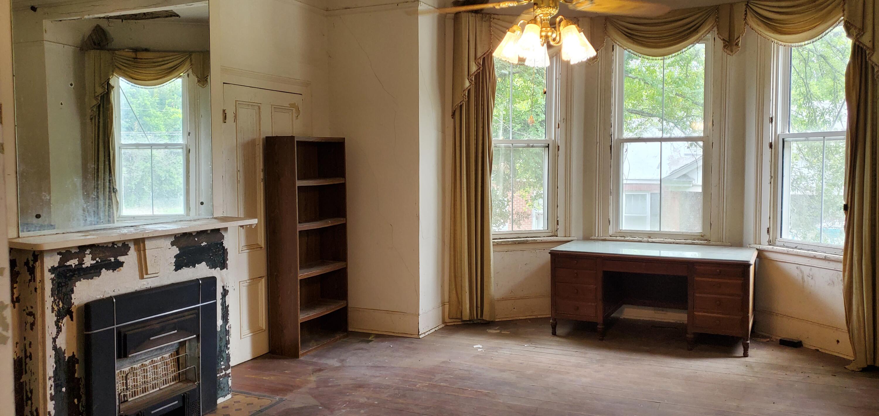 Worn living room with a peeling fireplace, tall bay windows, and a wooden desk.