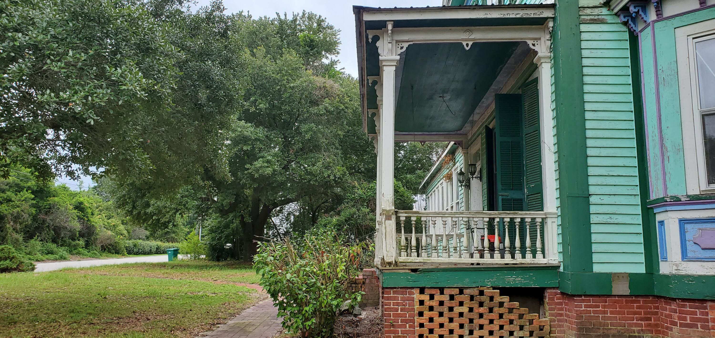 Mint-green house with a covered porch, ornate white railings, and mature trees beside the front walk.
