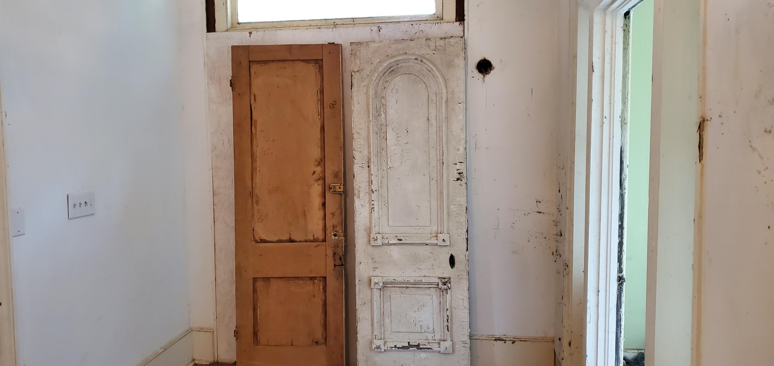 Vacant room with two weathered paneled doors leaning against the wall beside a window.
