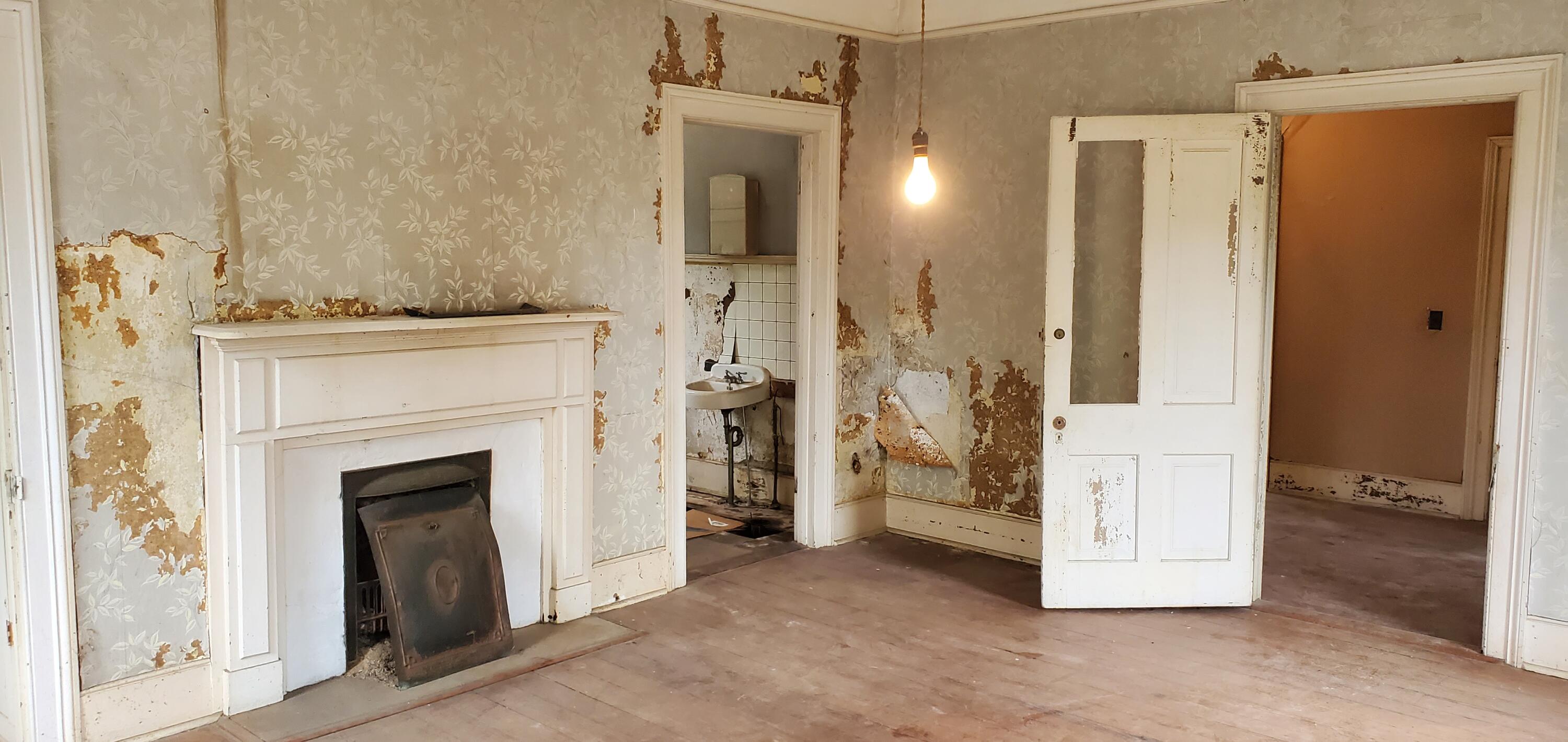 Vacant room with a white fireplace, peeling floral wallpaper, and a doorway to a small sink room.