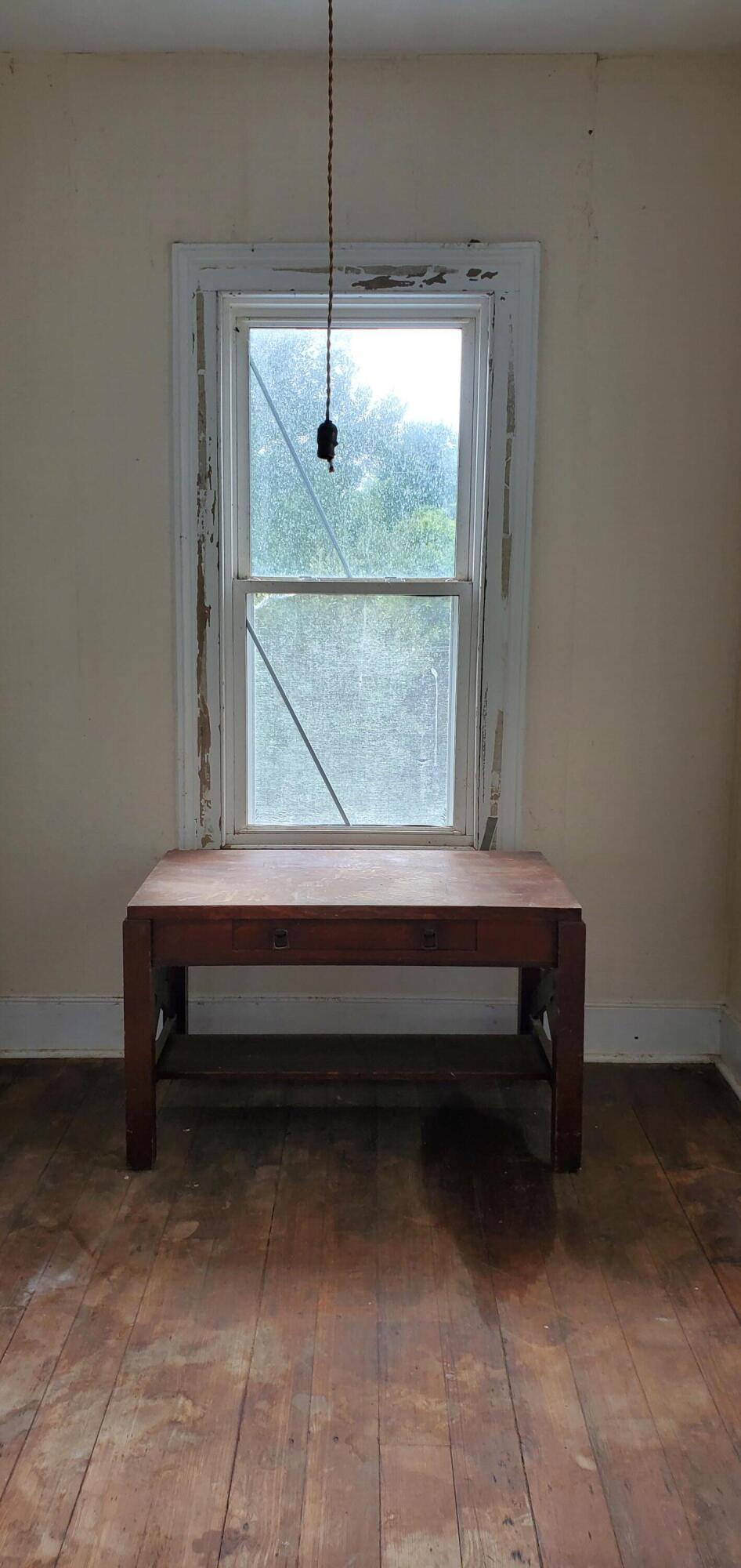 Wooden table beneath a tall window with peeling trim and worn plank floors.