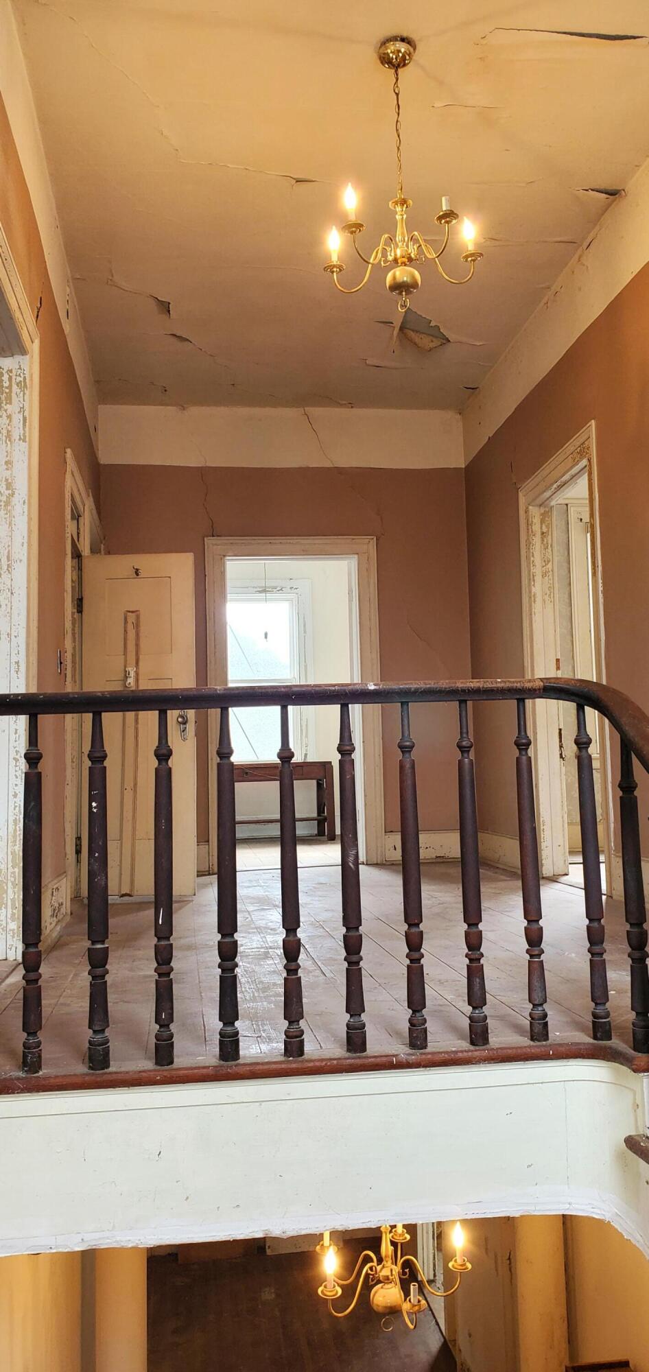 Vacant hallway with a wooden balustrade, brass chandelier, and extensively damaged ceiling.