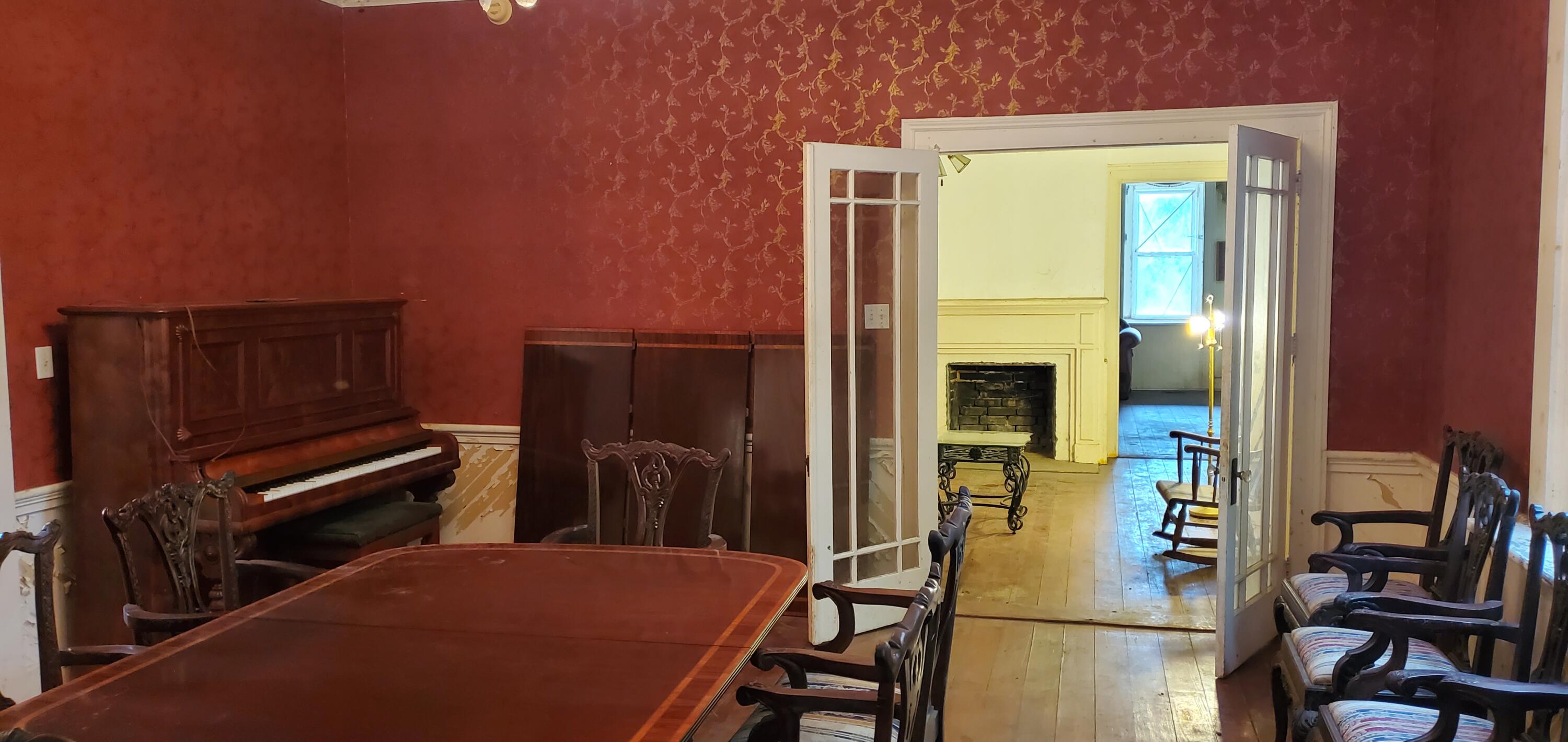 Dining room with red patterned walls, a wooden table, upright piano, and open doors to a fireplace room.