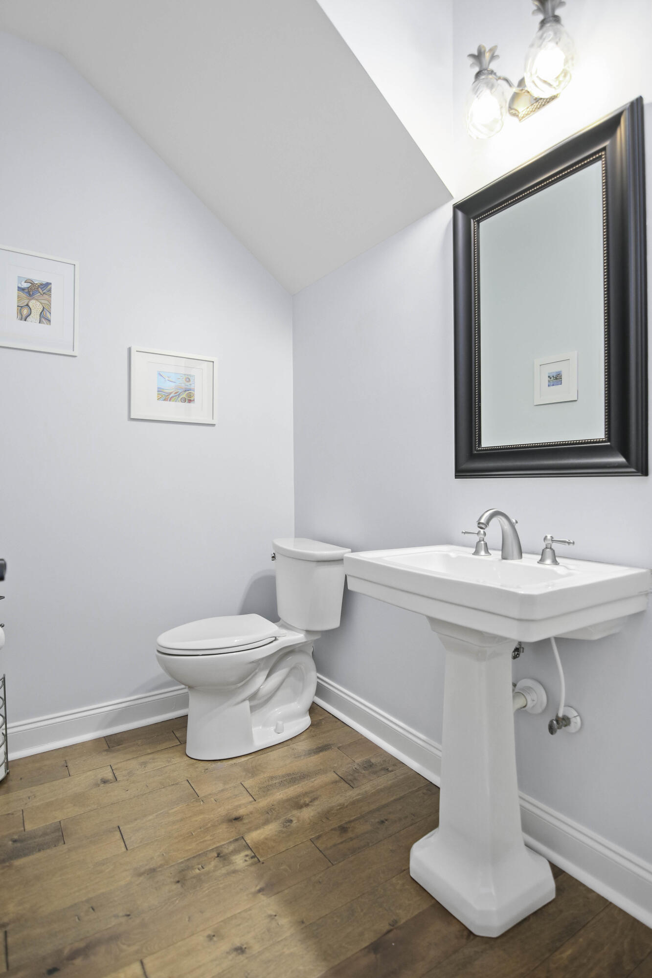 Bathroom with a pedestal sink, toilet, large framed mirror, and wood plank flooring.