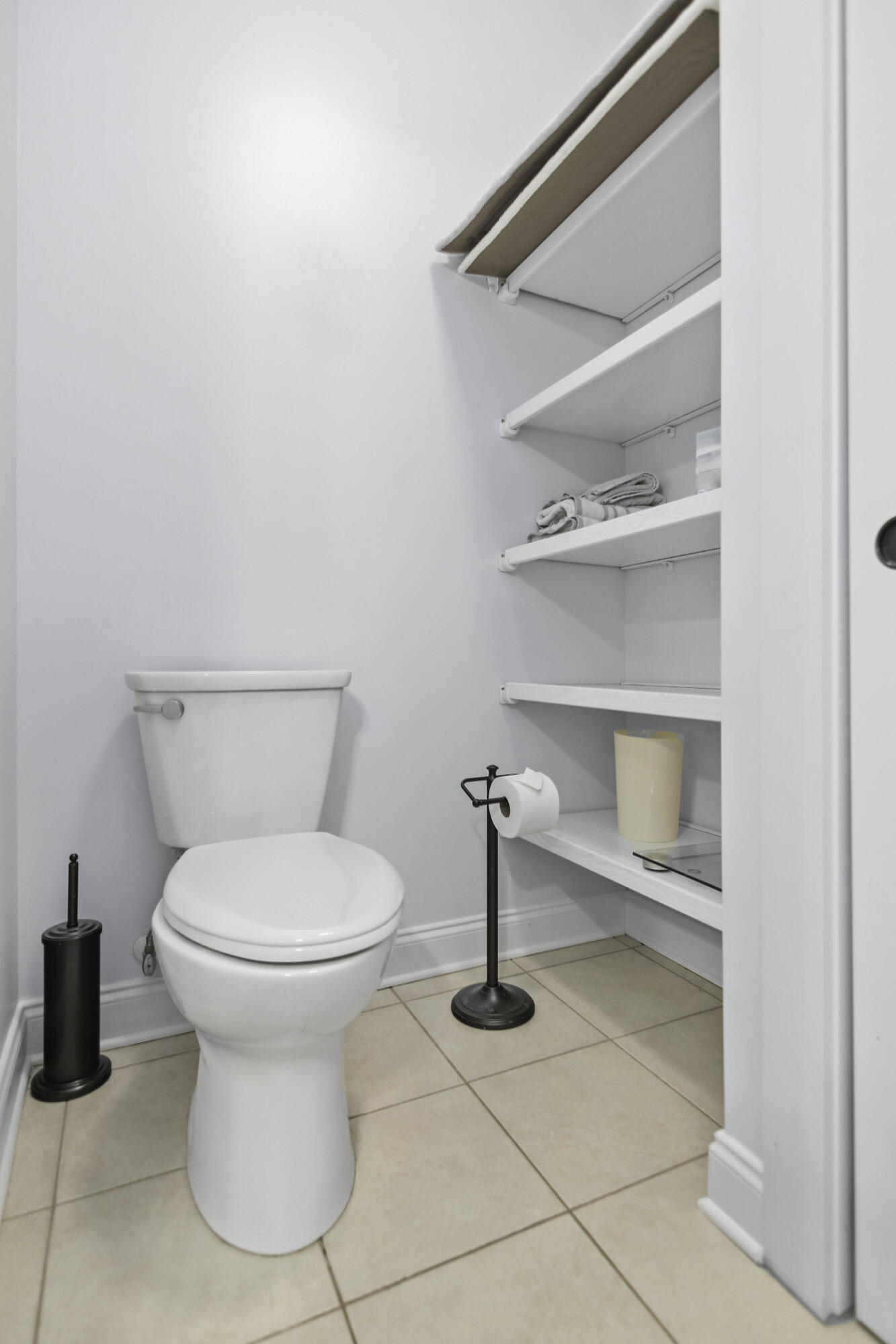 Bathroom with a white toilet, beige tile floor, and built-in shelves with folded towels.