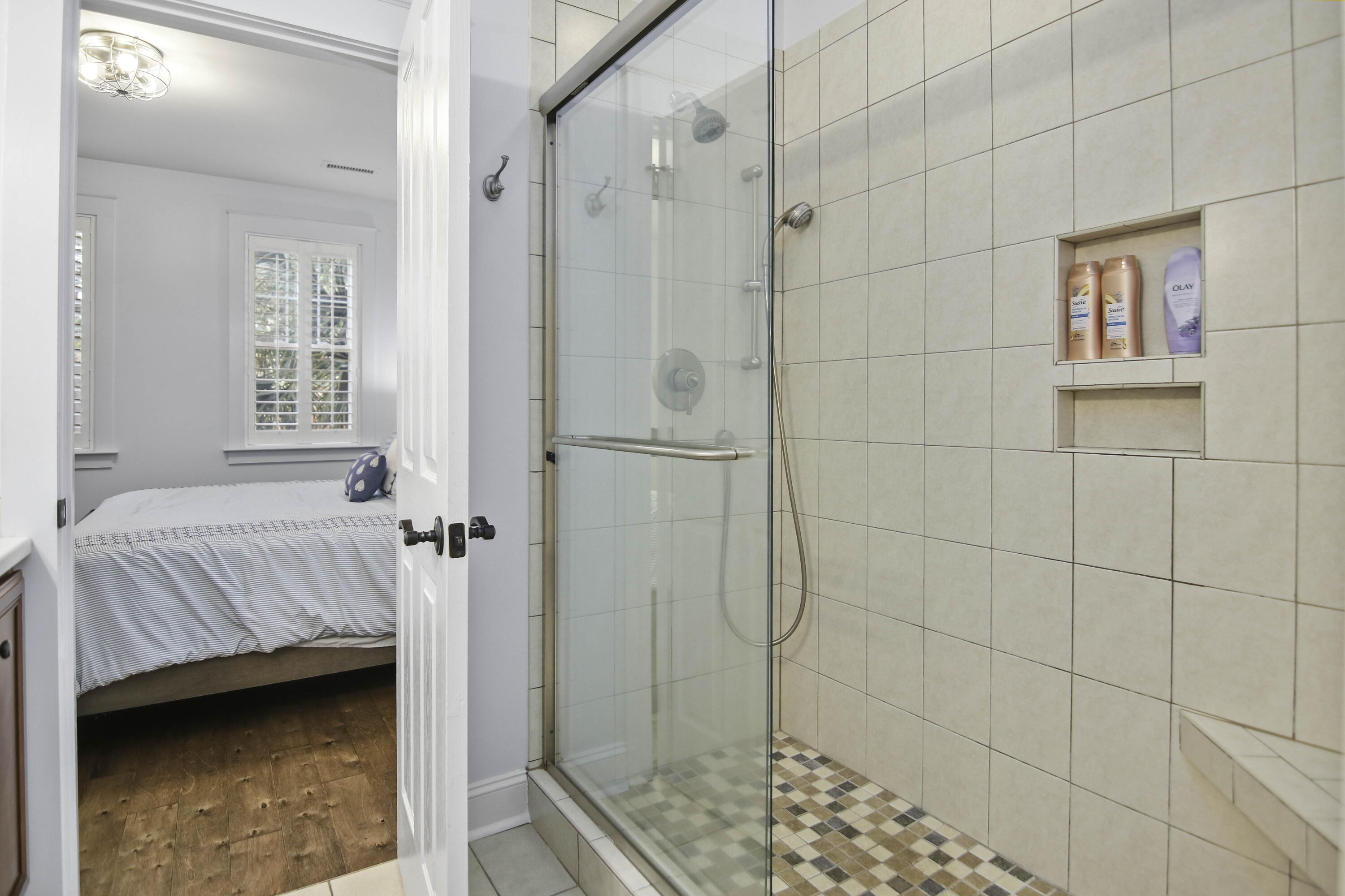 Tiled bathroom with a glass walk-in shower and a view into a bright bedroom.