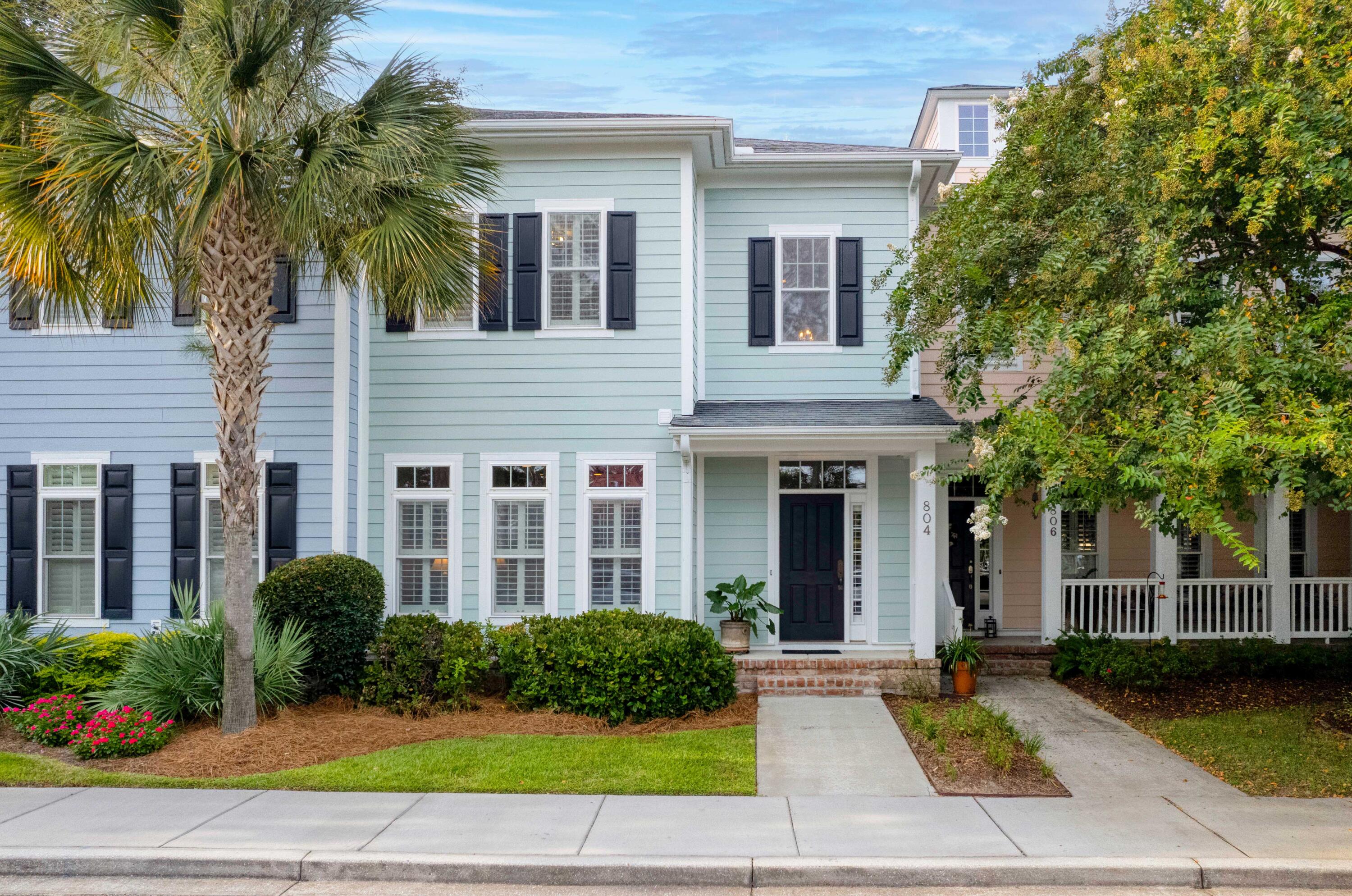 Pale blue two-story house with black shutters, brick entry steps, a palm tree, and lush landscaping.