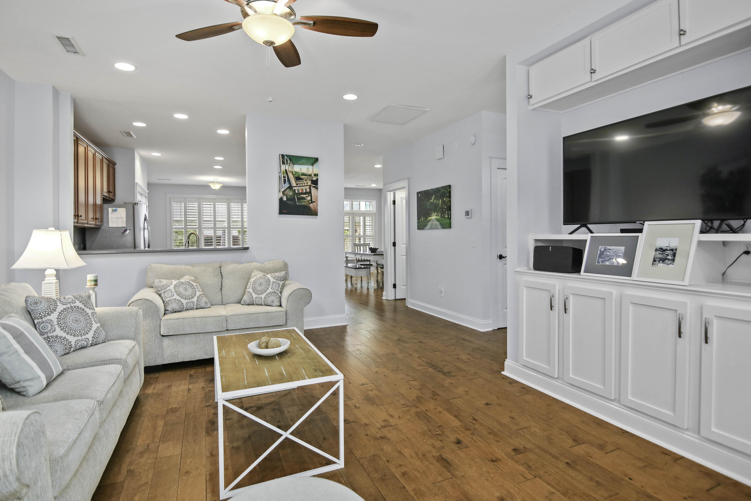 Living room with built-in TV cabinetry, light sofas, hardwood floors, and an open view toward the kitchen.