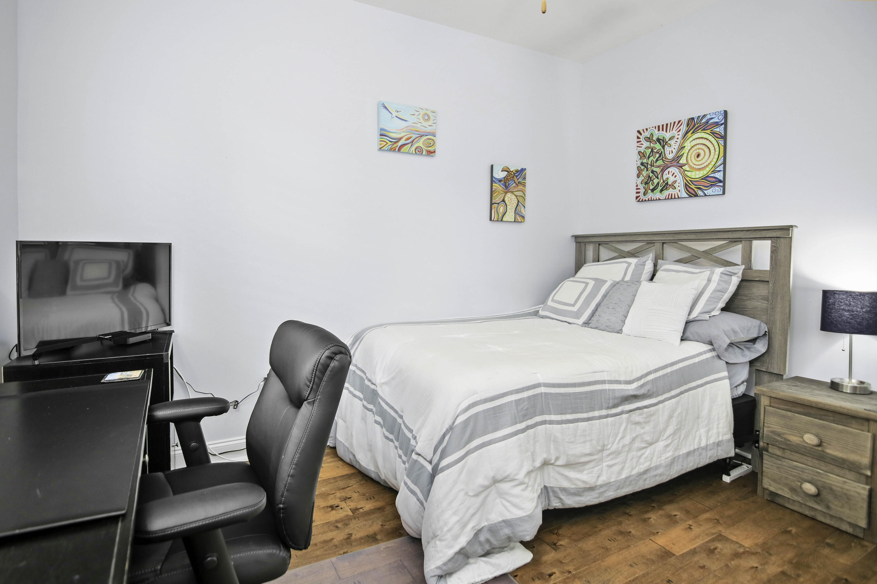 Bedroom with gray-and-white striped bedding, colorful wall art, hardwood floors, and a desk with an office chair.