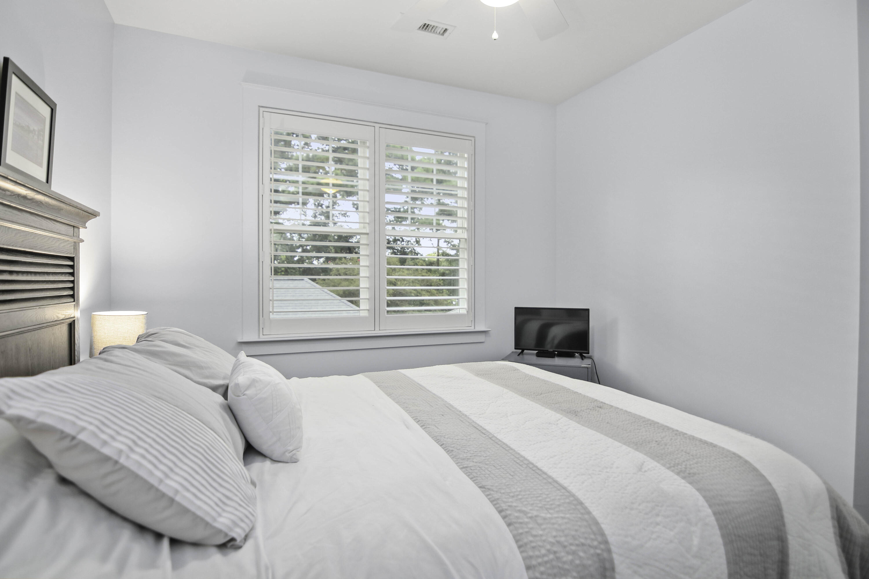 Furnished bedroom with a white bed, plantation-shuttered window, dark dresser, and small television.