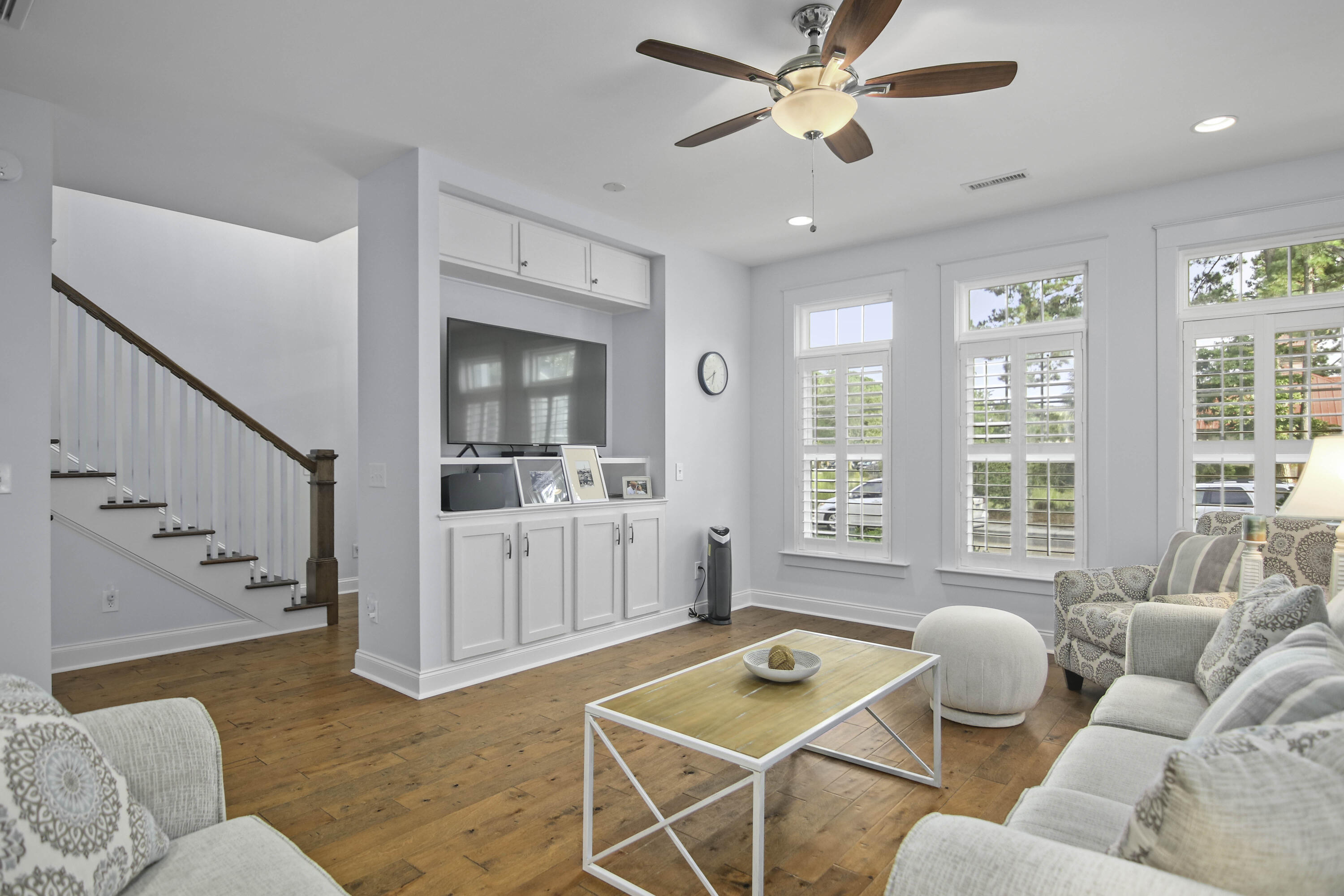 Furnished living room with hardwood floors, built-in media cabinets, shuttered windows, and a staircase.