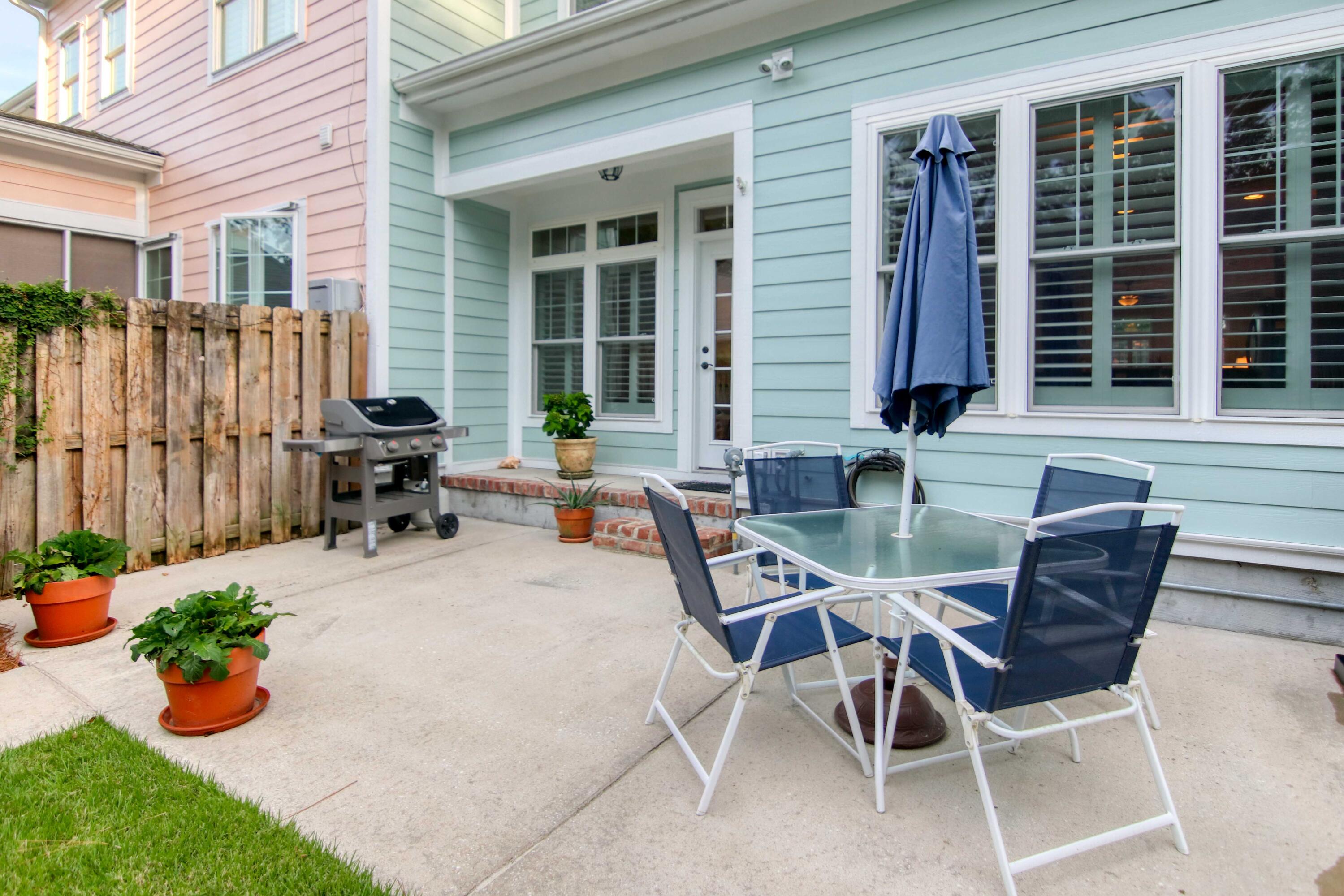 Backyard patio with a glass-top dining set, blue umbrella, gas grill, and potted plants beside a light blue home.