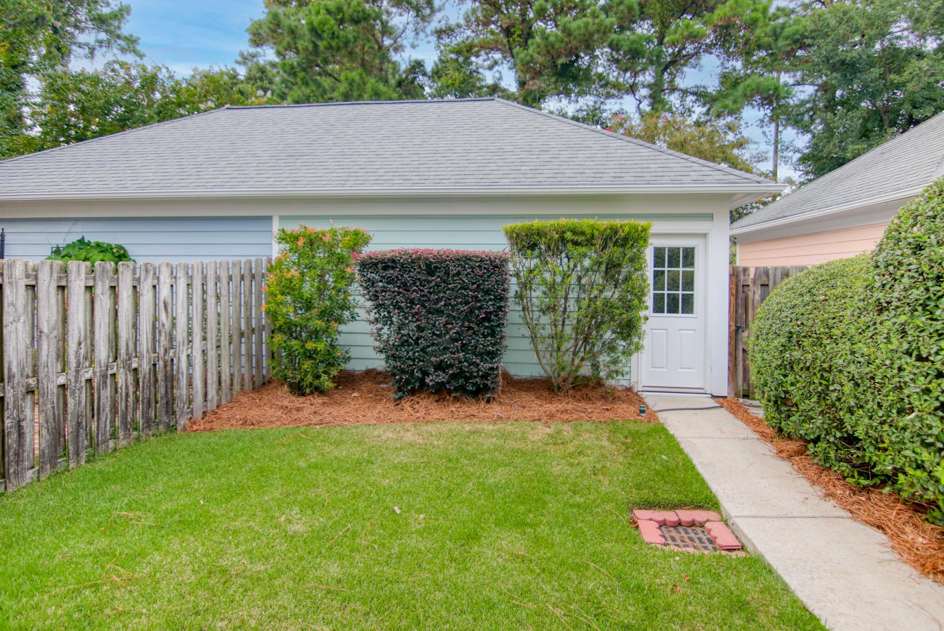 Light blue home’s side entry with white door, trimmed shrubs, wooden fence, and lawn.