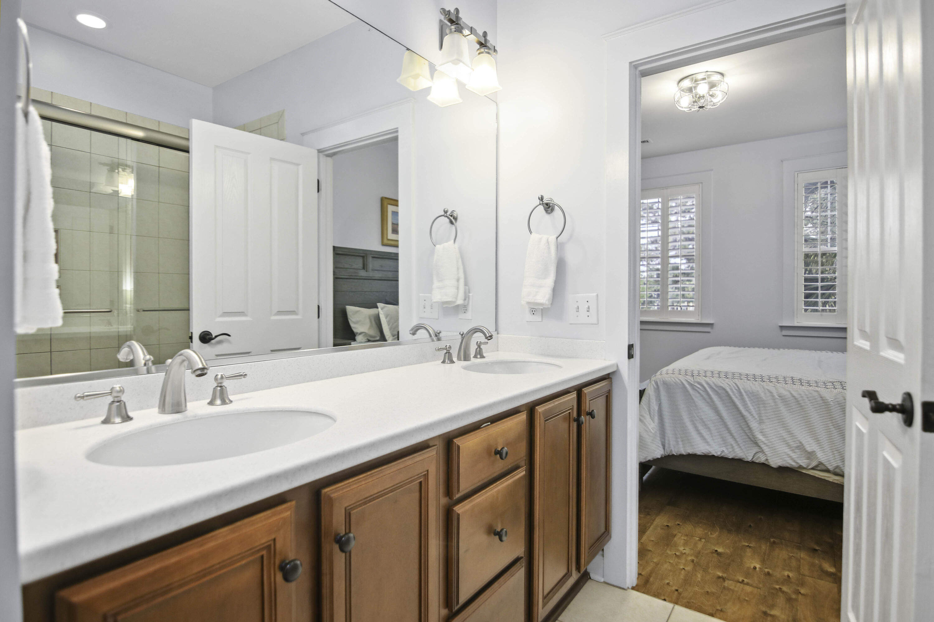 Bathroom with a double-sink vanity and glass shower, opening to a bedroom with shuttered windows.