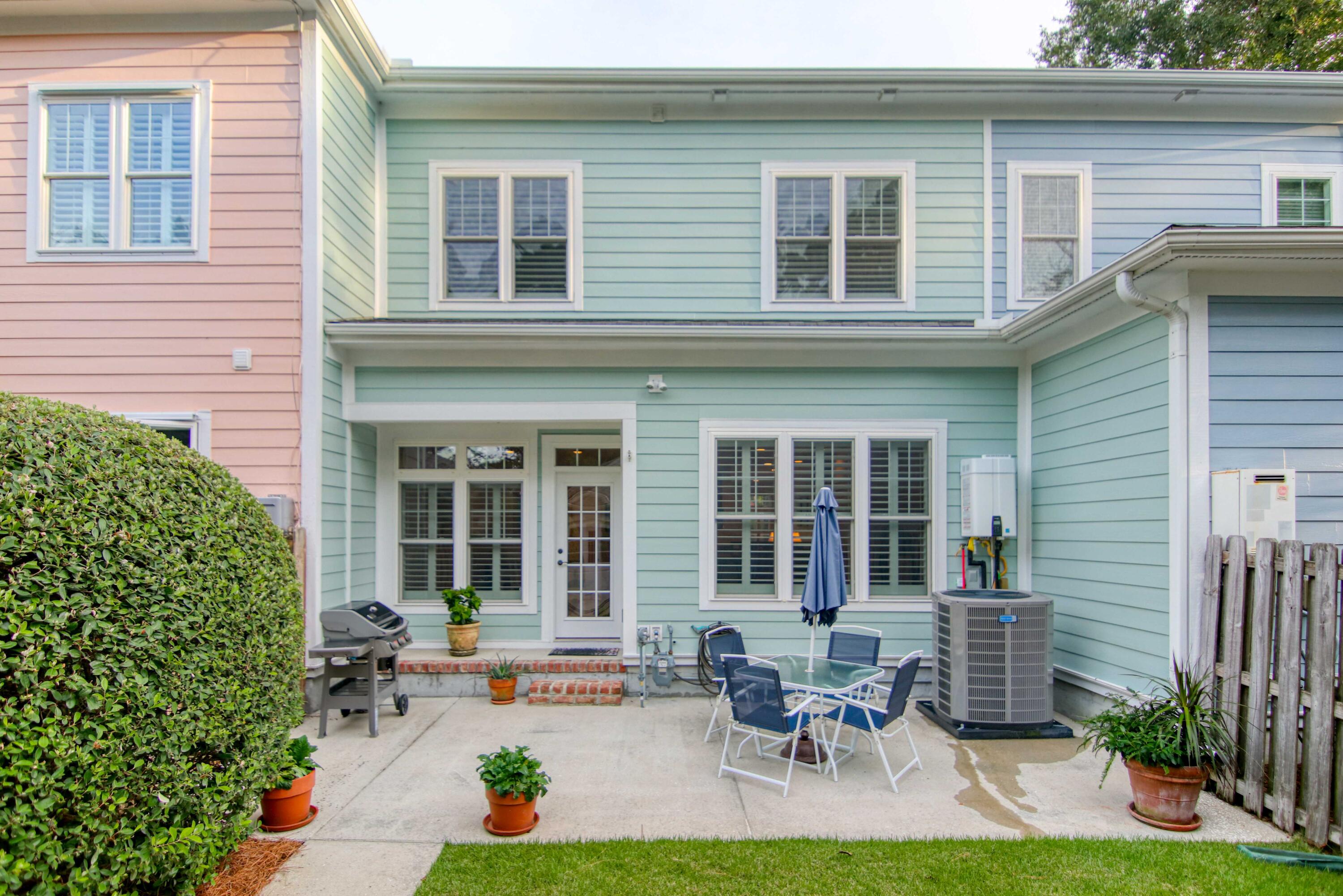 Back patio with outdoor dining set, closed umbrella, potted plants, and grill beside a pale blue home.