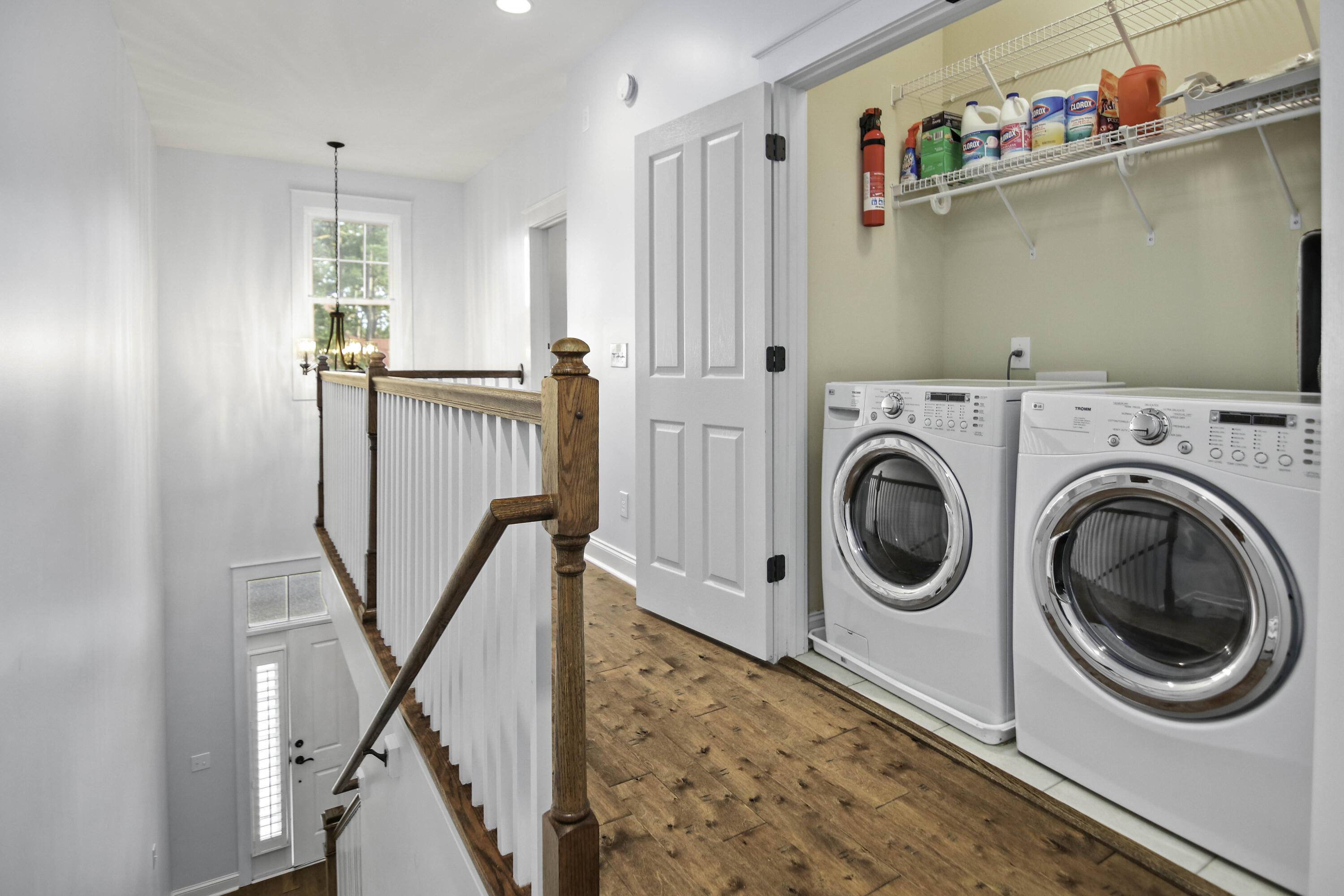 Upstairs laundry area with two front-loading machines, stocked wire shelves and a wood-floored stair landing.