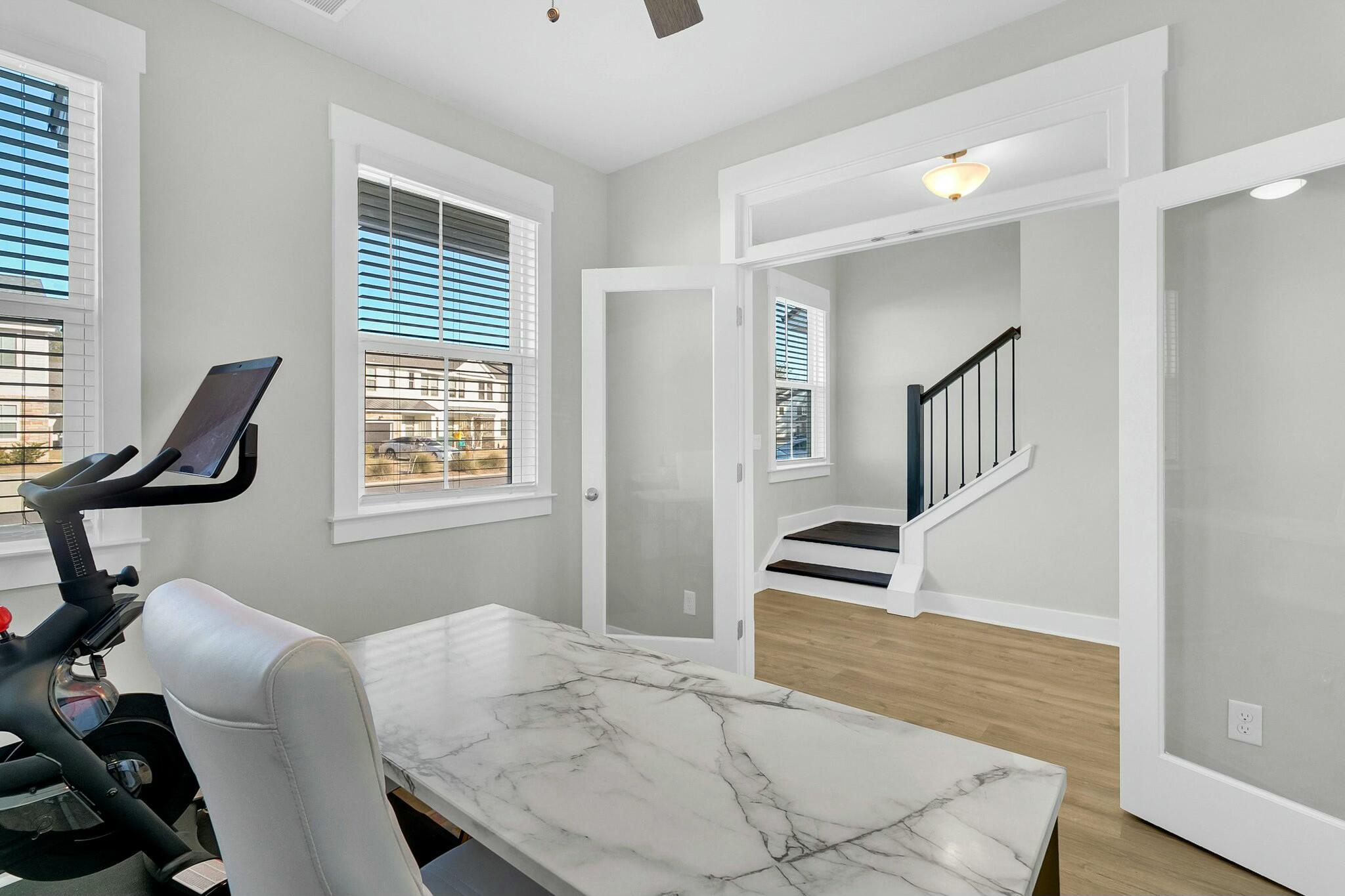 Home office with a marble-patterned desk, exercise bike, shuttered windows, and view of the staircase.