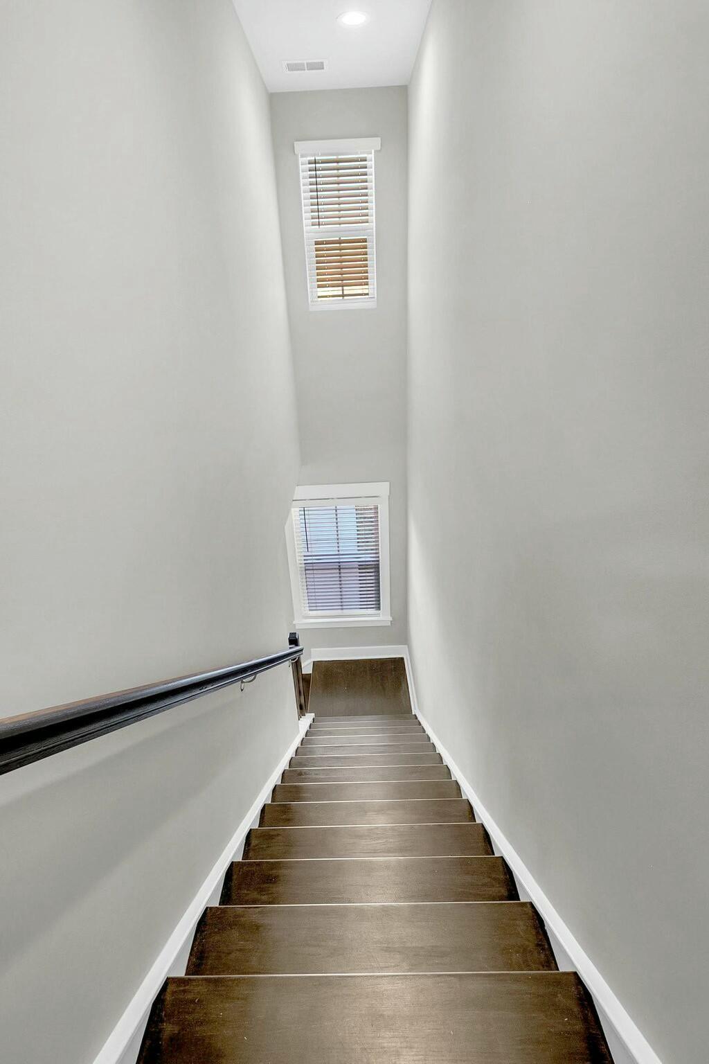 Tall stairwell with dark wood steps, a dark handrail, and two windows with blinds.