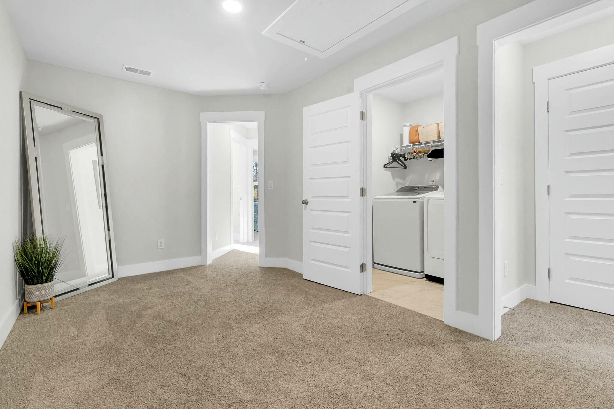 Carpeted bedroom with a leaning full-length mirror, potted plant, and doorway to a laundry area.