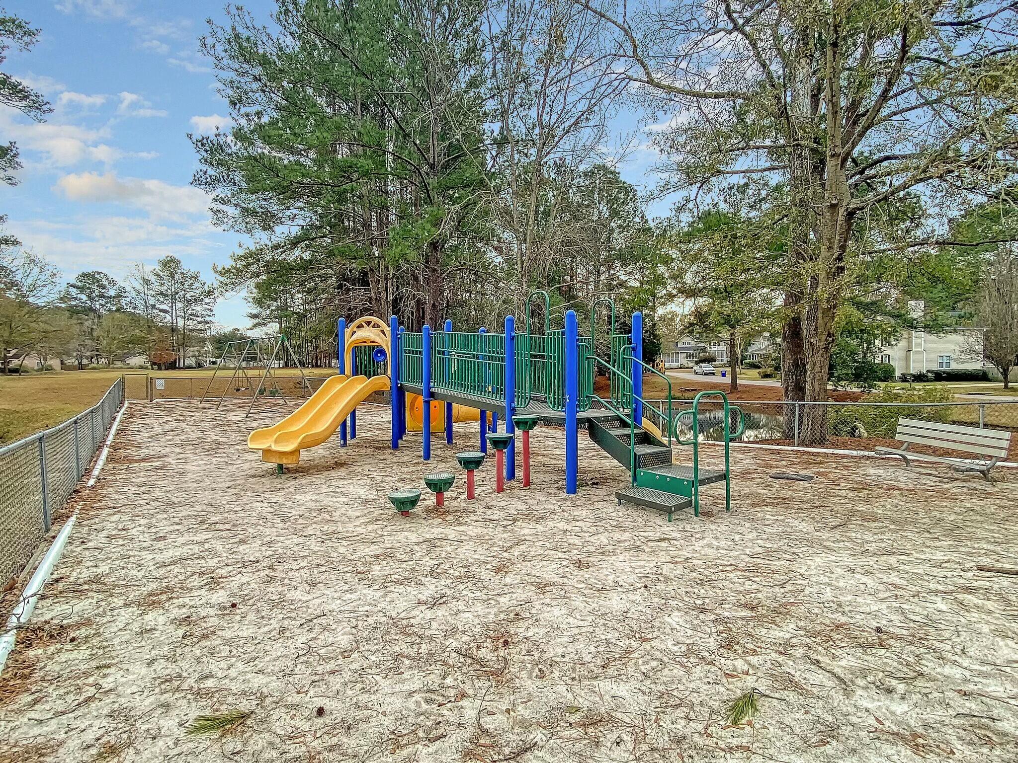 Fenced playground with yellow slides, a colorful climbing structure, and a bench beneath mature trees.