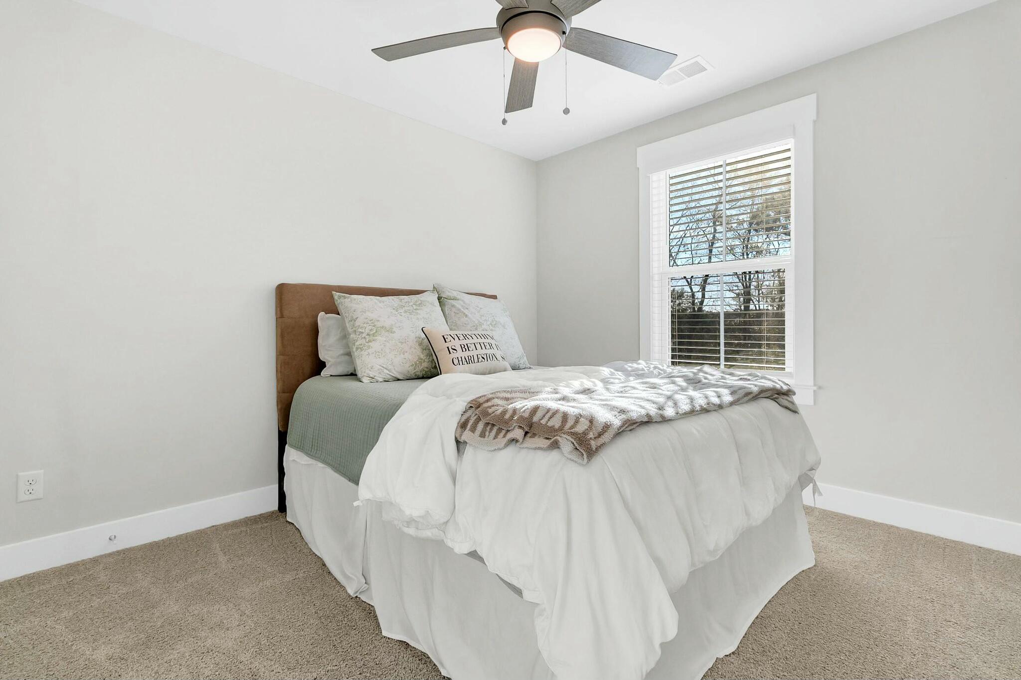 Staged bedroom with a white bed, tan upholstered headboard, and large window with blinds.