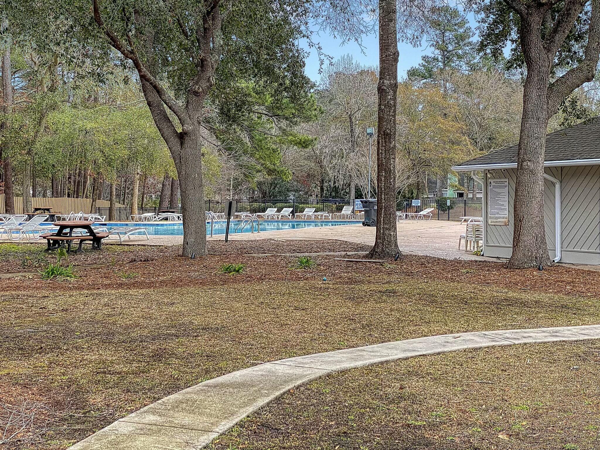 Community pool with white lounge chairs beneath mature trees and a curved sidewalk in the foreground.