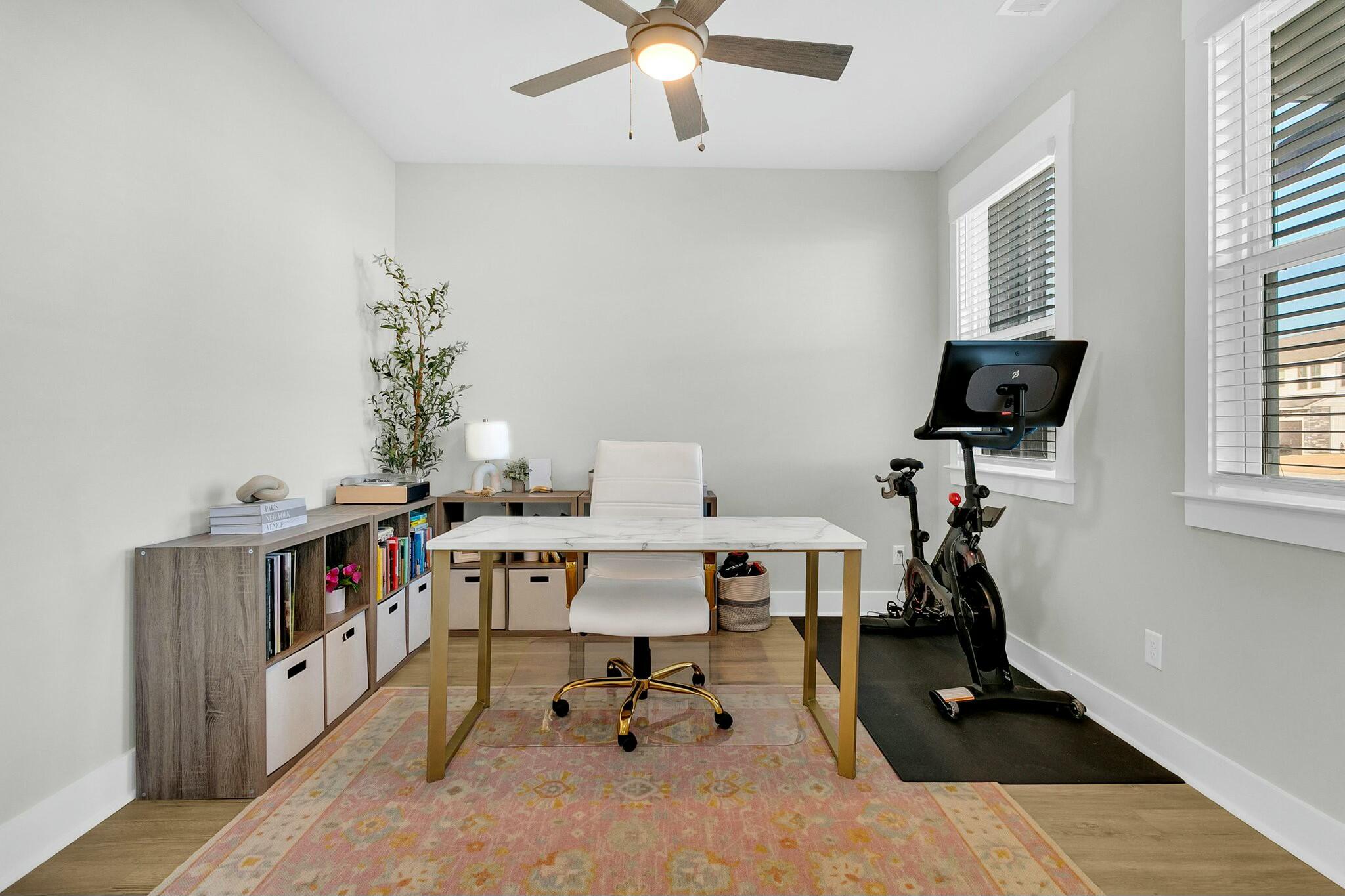 Home office with a white desk, bookcase, and stationary exercise bike by the windows.