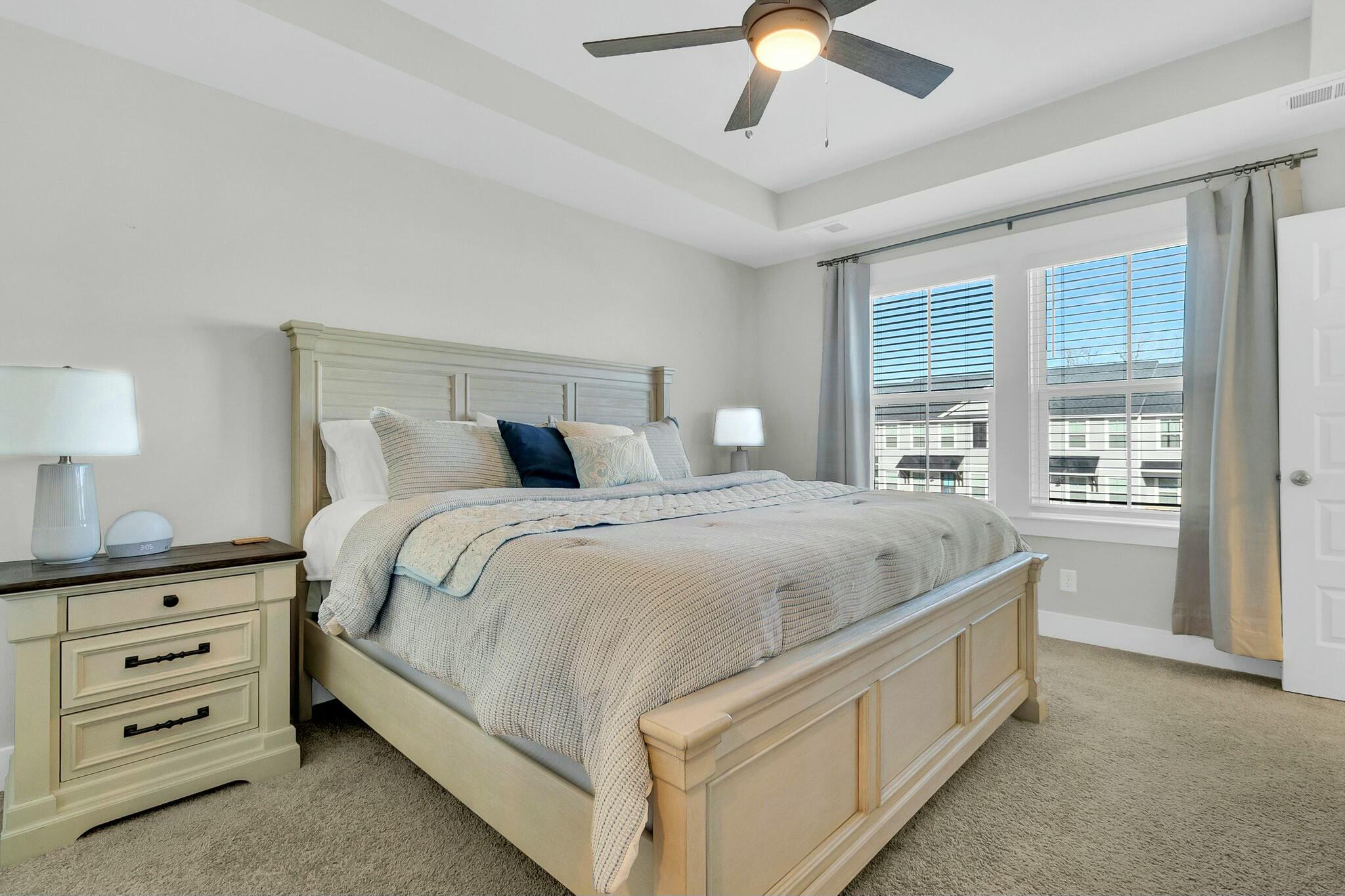 Staged bedroom with a large cream bed, tray ceiling, ceiling fan, and two bright windows.
