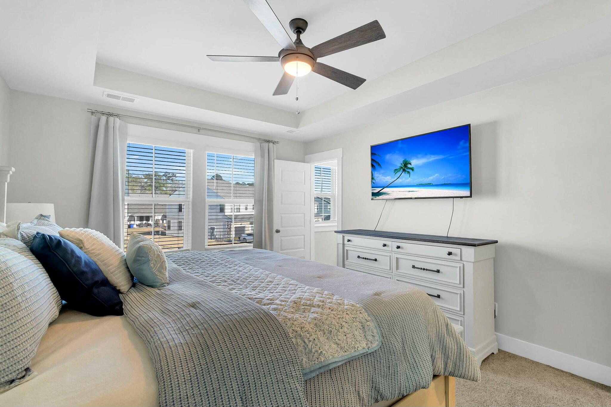 Furnished bedroom with a gray quilted bed, white dresser, and windows with blinds.