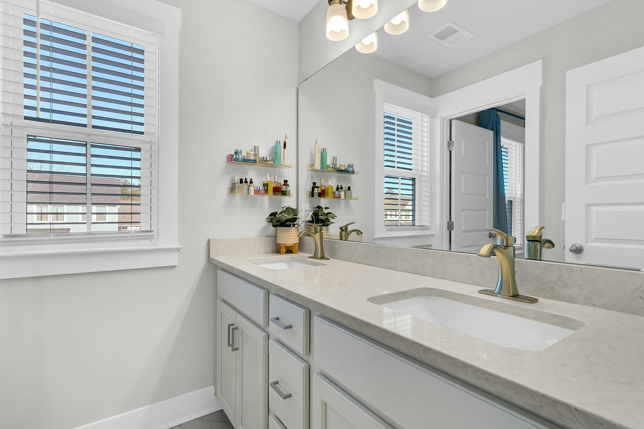 Bright bathroom with a double-sink vanity, large mirror, brass-tone faucets, and windows with white blinds.