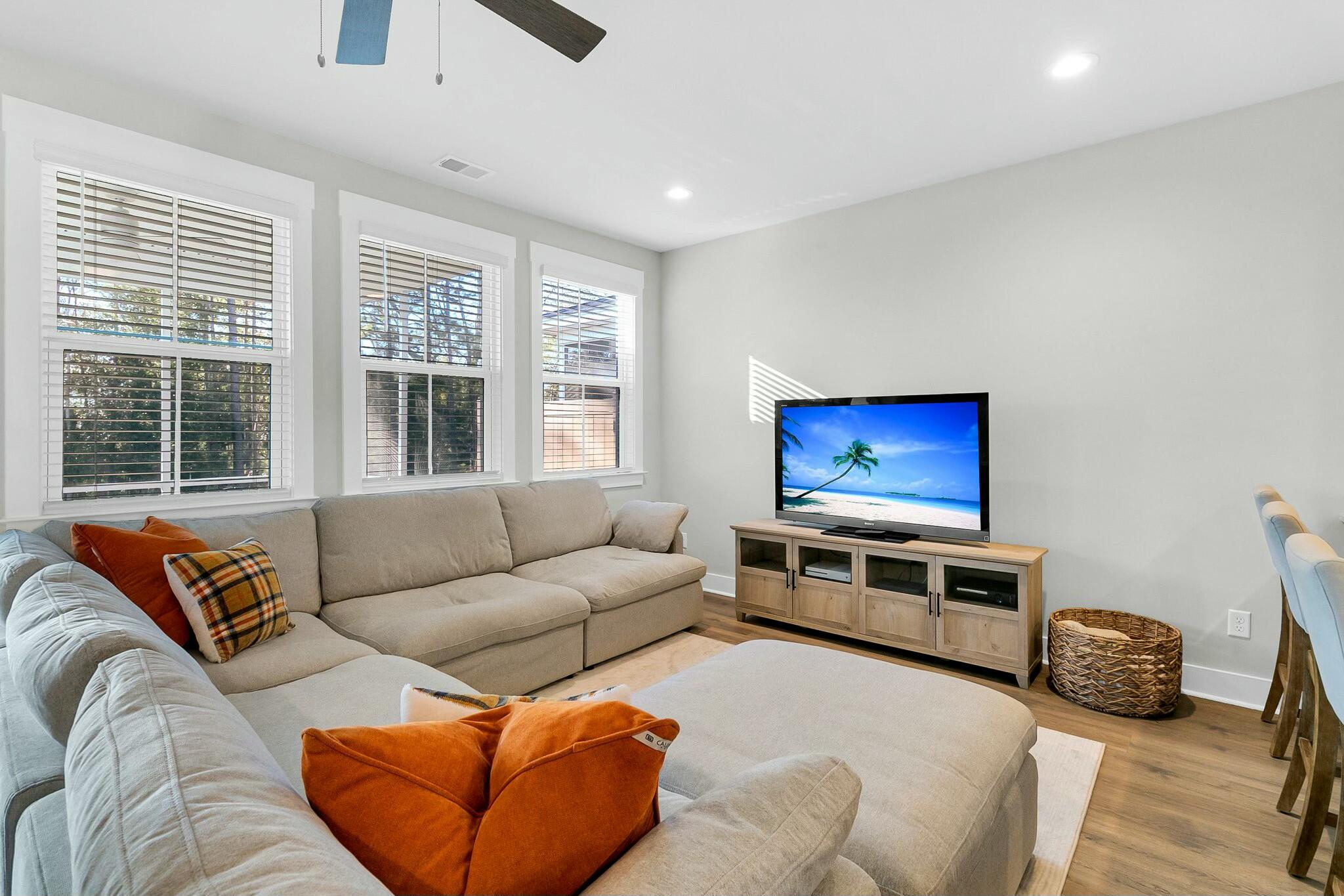 Living room with a beige sectional, orange pillows, three shuttered windows, and a television.
