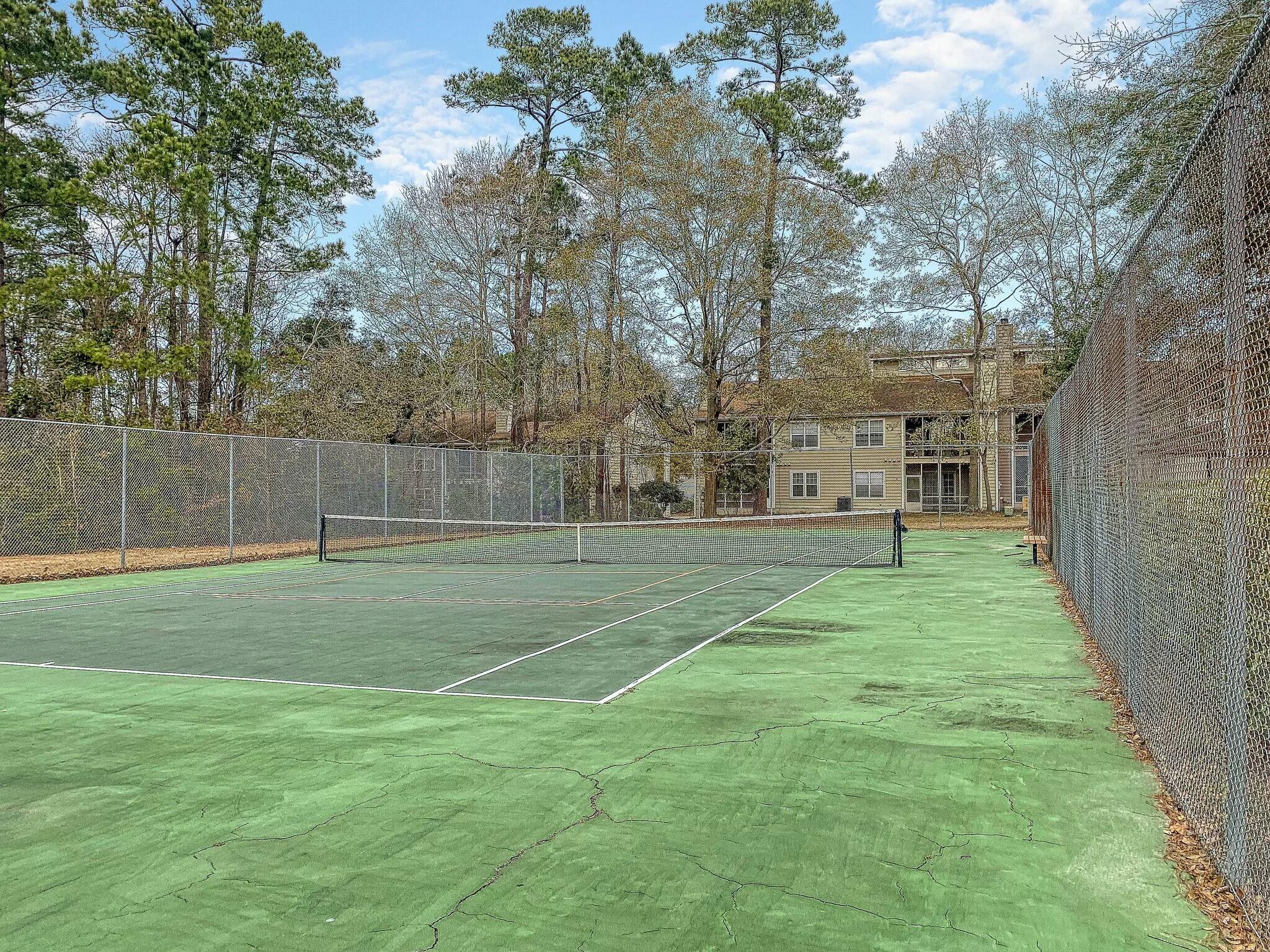 Fenced outdoor tennis court with a cracked green surface and residential building beyond.