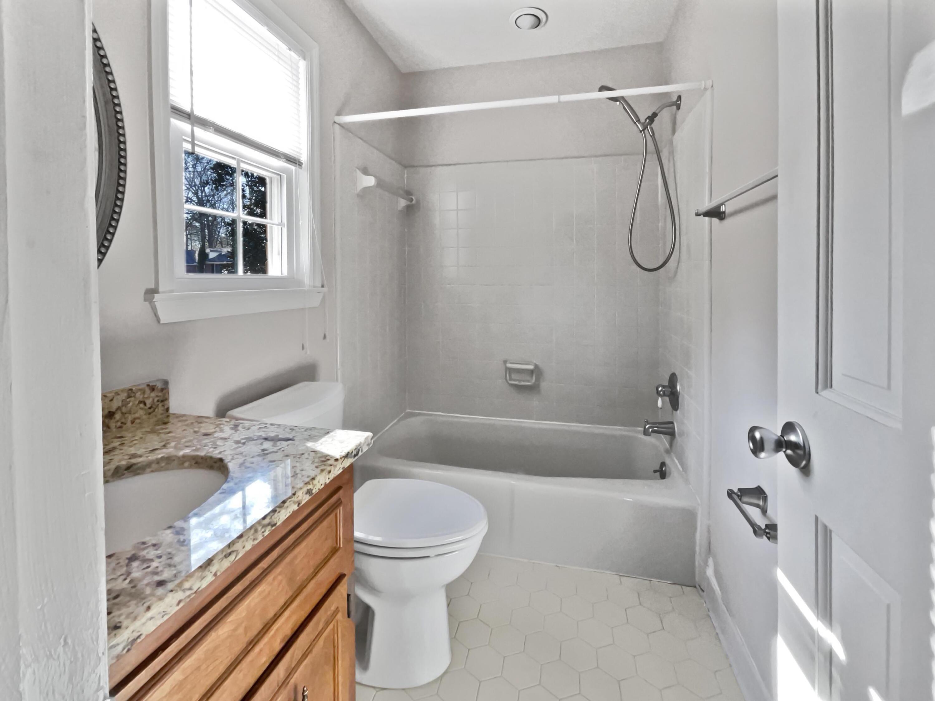 Bathroom with a granite-topped wood vanity, tub and shower, and white hexagonal floor tile.