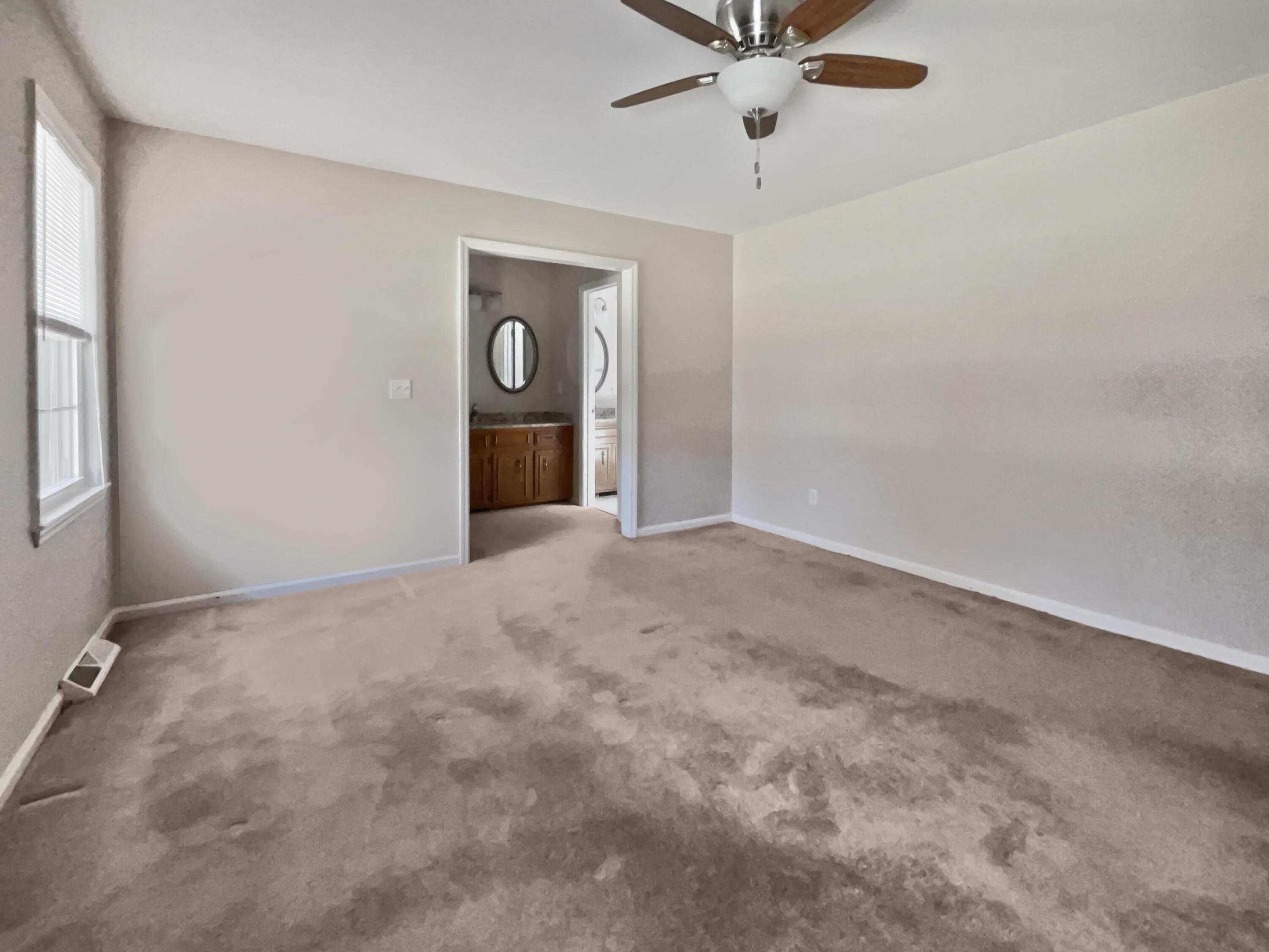 Empty bedroom with a ceiling fan, marked carpet, and doorway to a bathroom with a wood vanity.