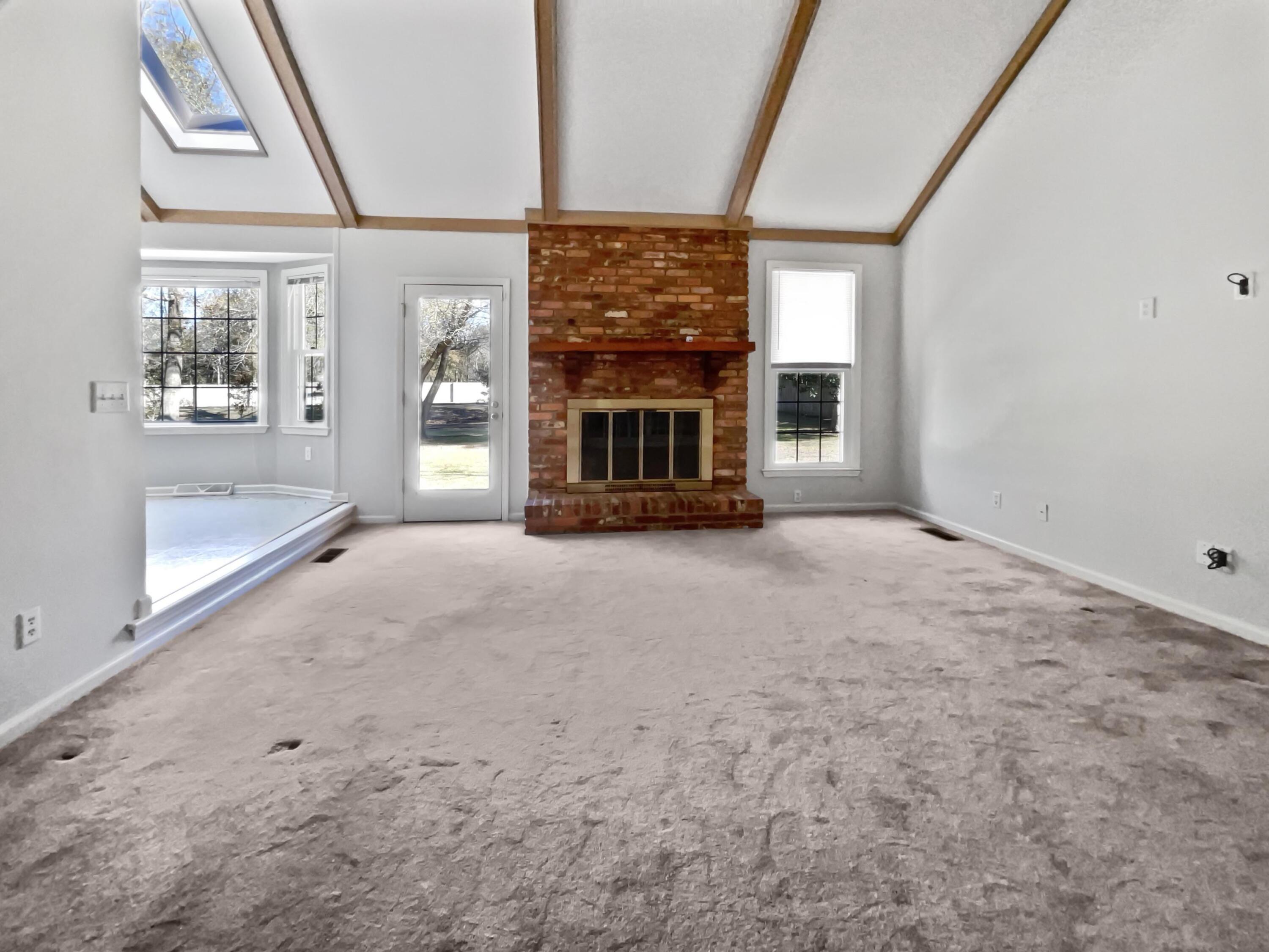Vacant living room with a brick fireplace, vaulted beamed ceiling, skylight, and worn carpet.