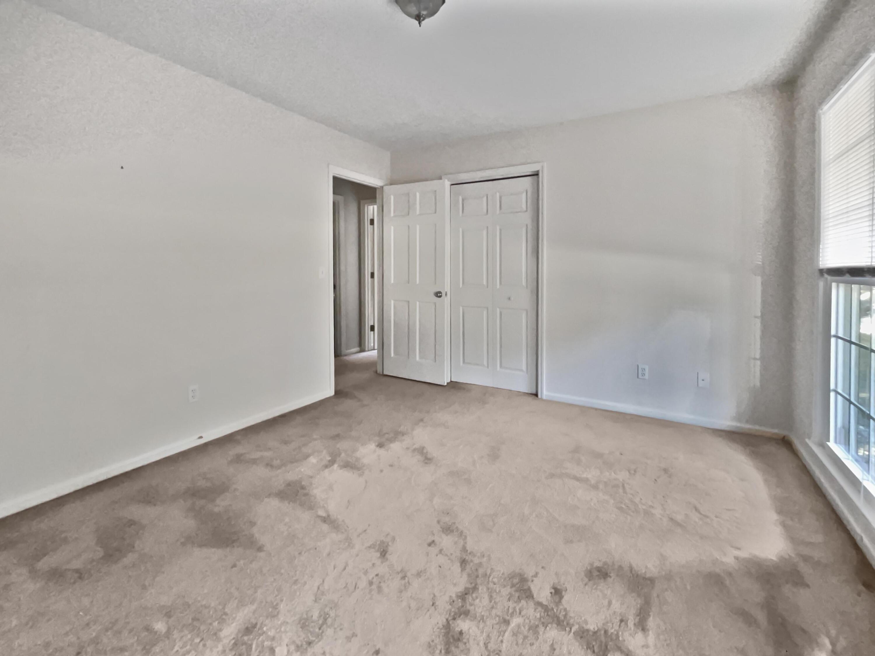 Vacant bedroom with stained beige carpet, open closet doors, and a window with blinds.