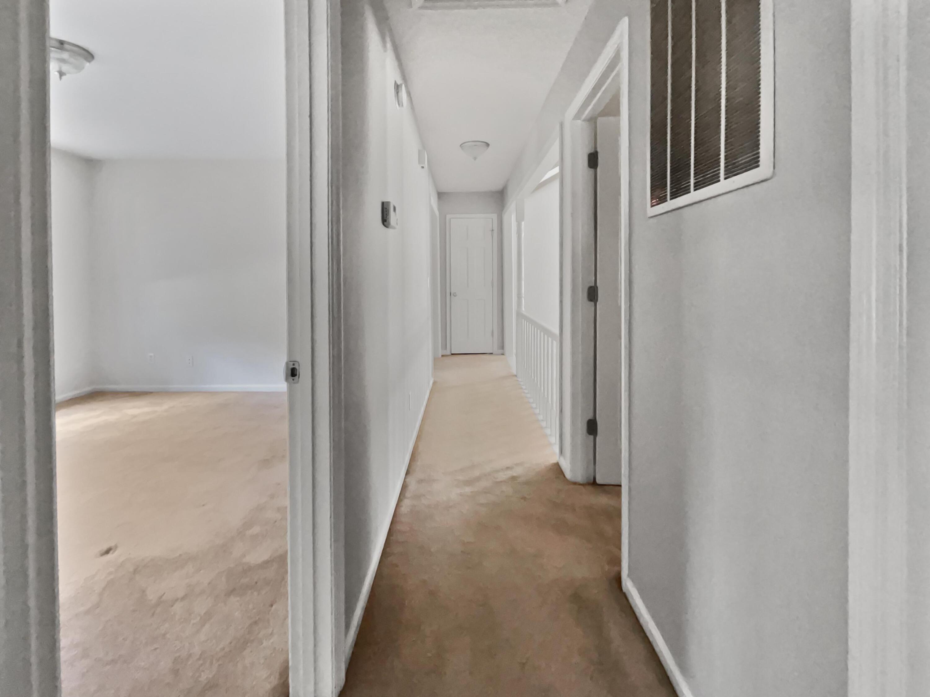 Empty carpeted hallway with white walls and a doorway to an unfurnished room.