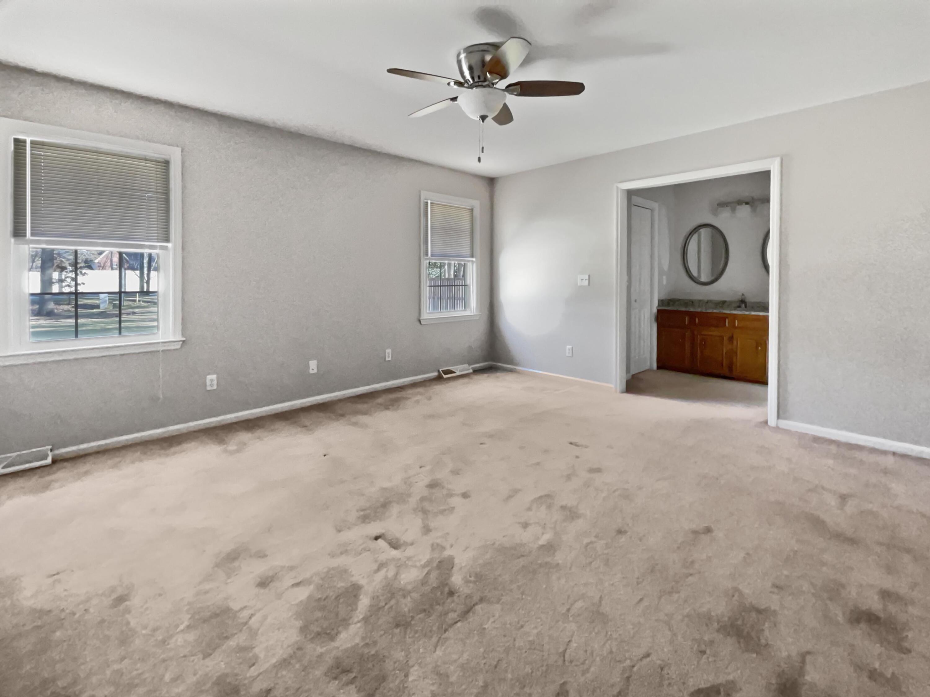 Empty carpeted bedroom with two windows, a ceiling fan, and a bathroom vanity visible through the doorway.