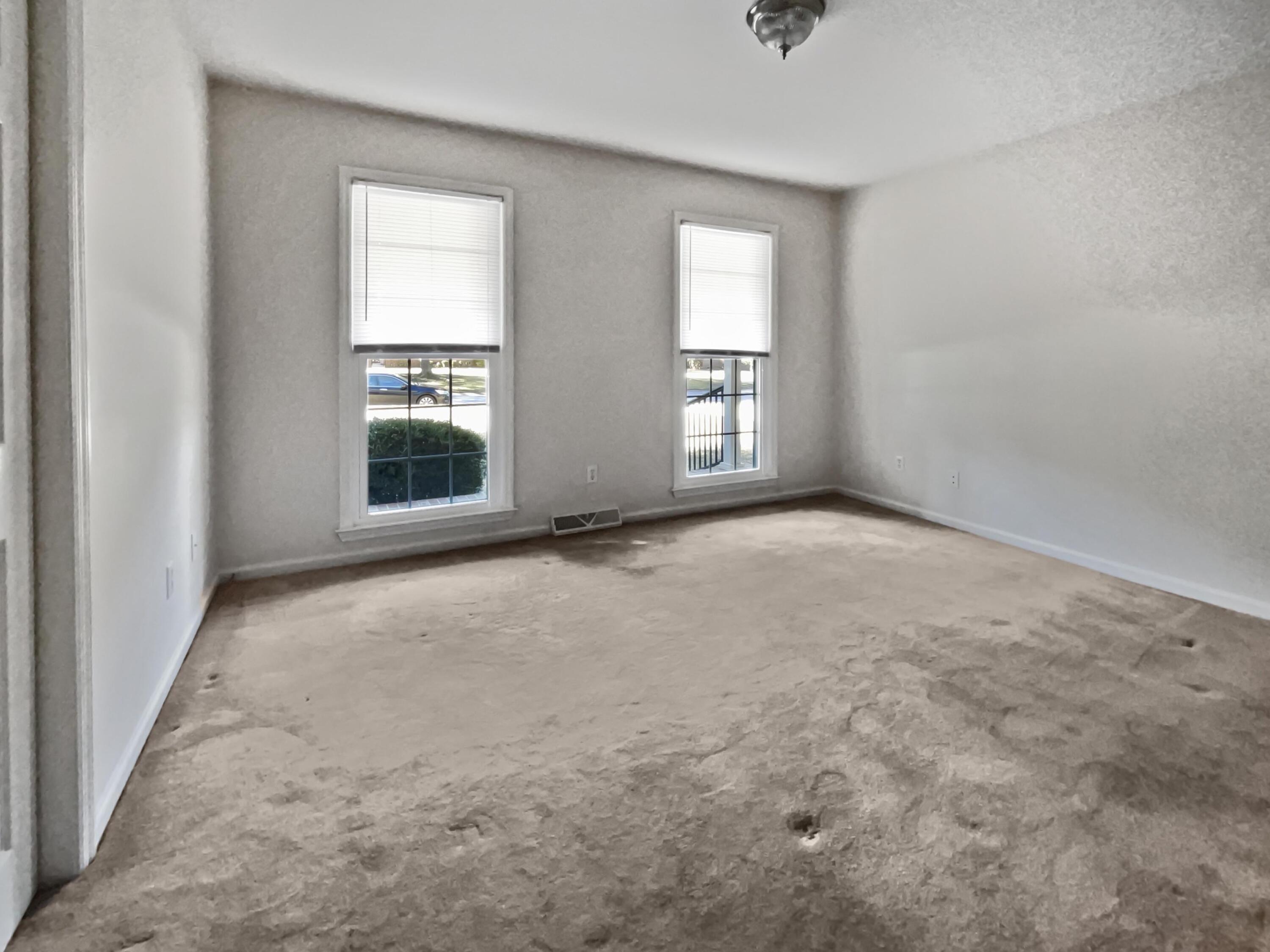 Empty room with two windows and worn, stained beige carpet.