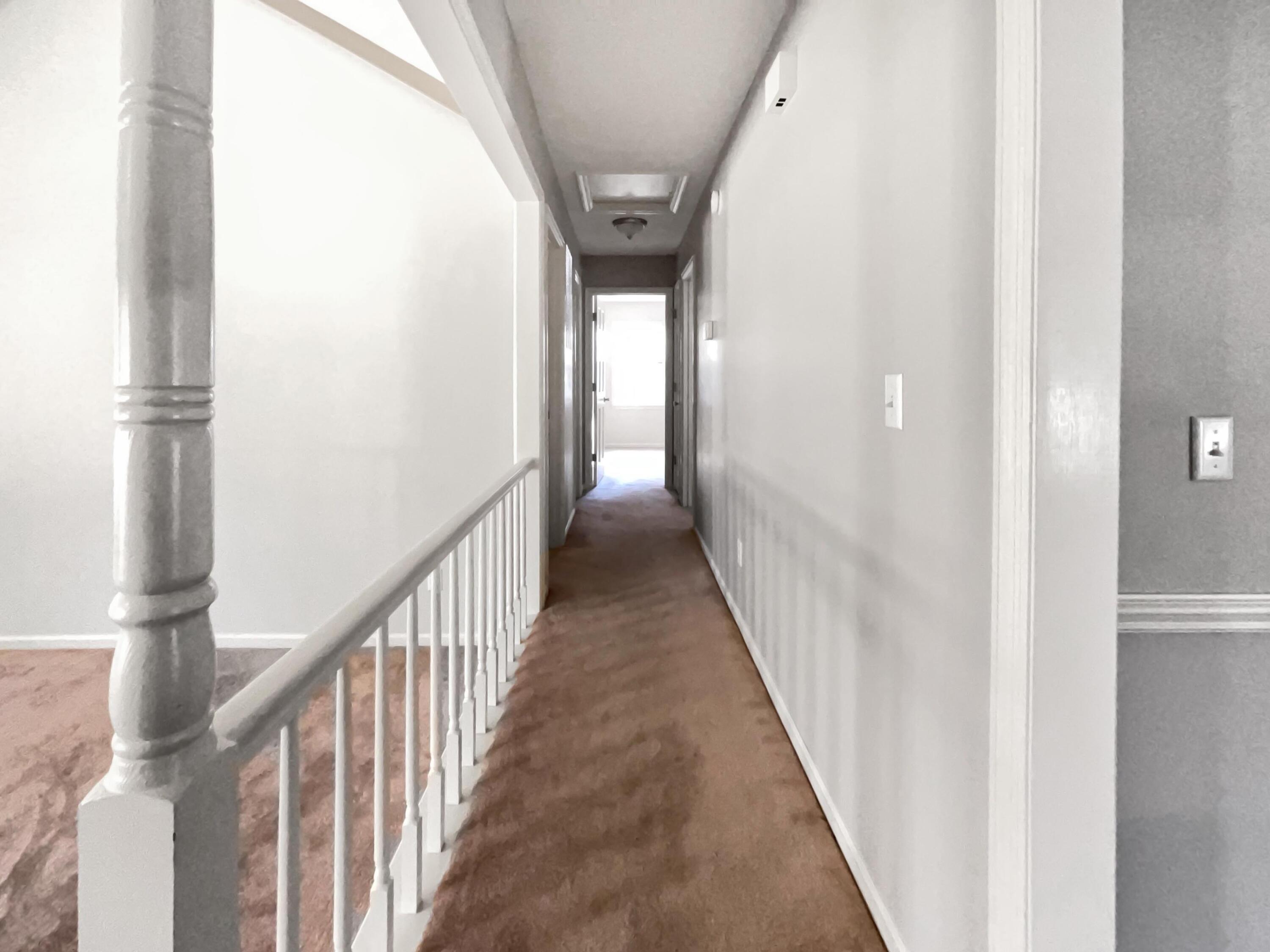 Upstairs hallway with brown carpet, white stair railing, and a bright doorway at the far end.