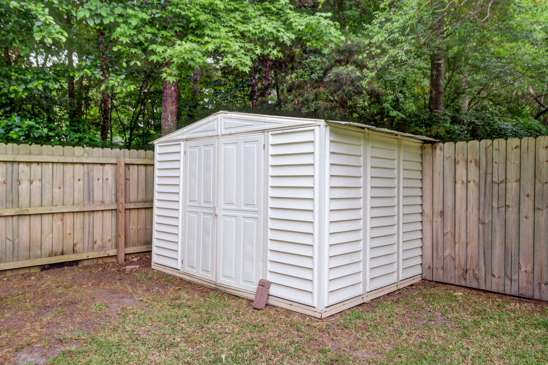 White storage shed with double doors in a fenced, tree-shaded backyard.