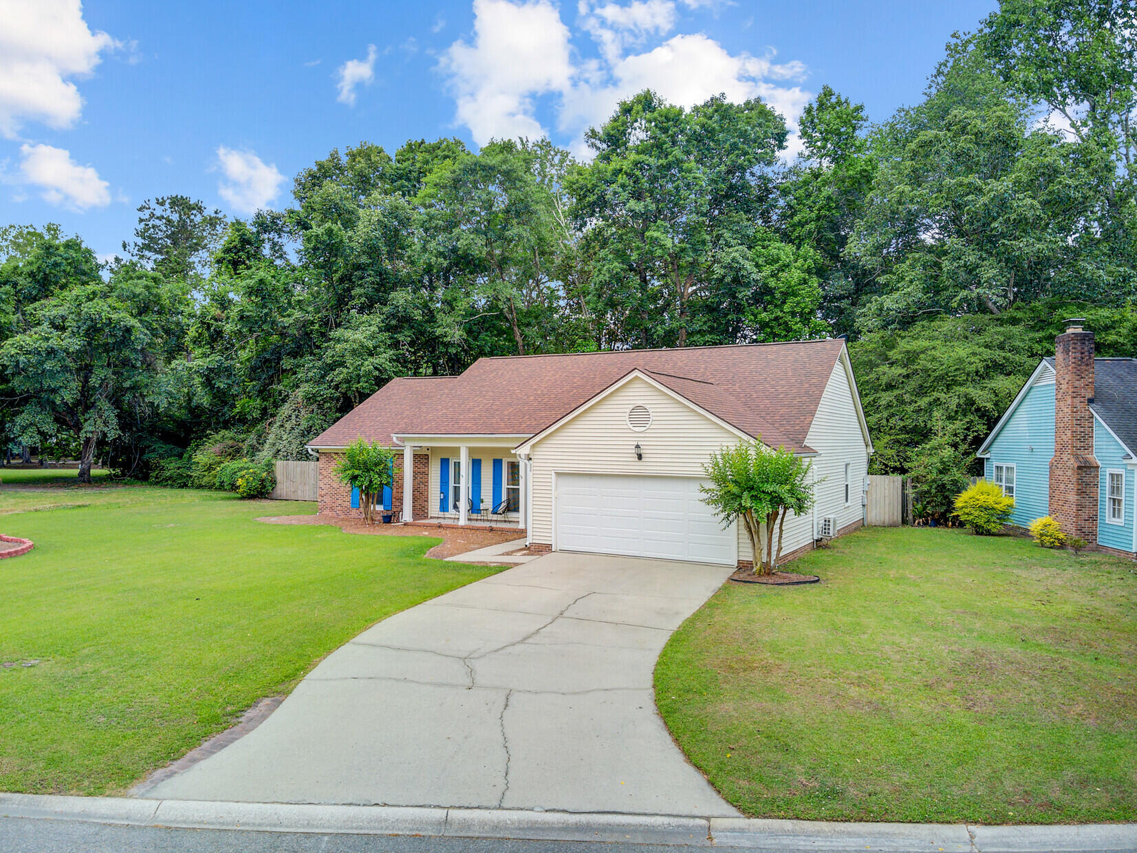 Single-story beige house with an attached garage, curved driveway, and tree-lined yard.
