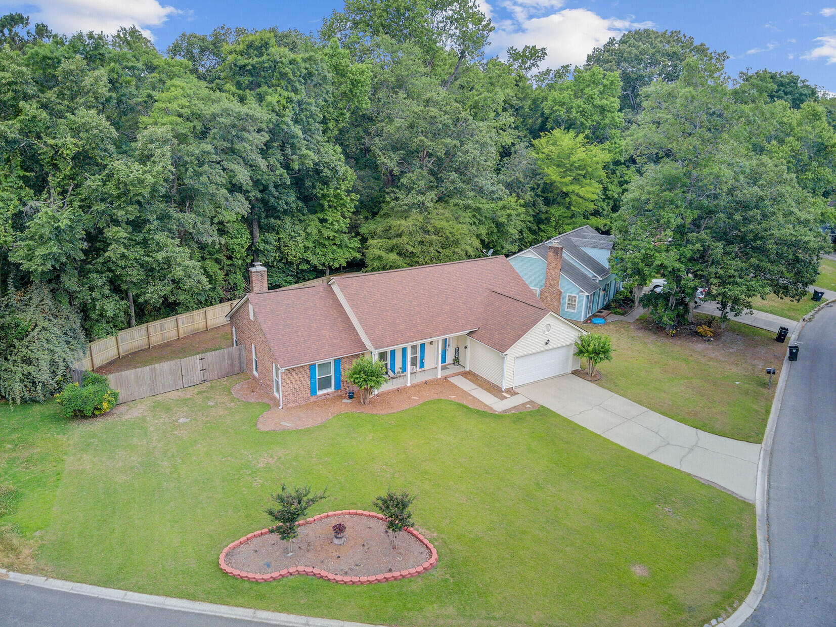 Aerial view of a brick house with blue shutters, a broad lawn, and dense woods behind it.